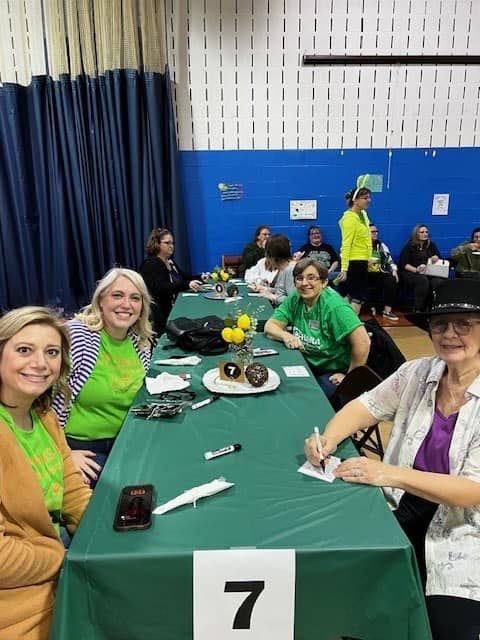 People seated at a table with a green tablecloth, numbered 7. Some are smiling and writing. Yellow flowers in the center.