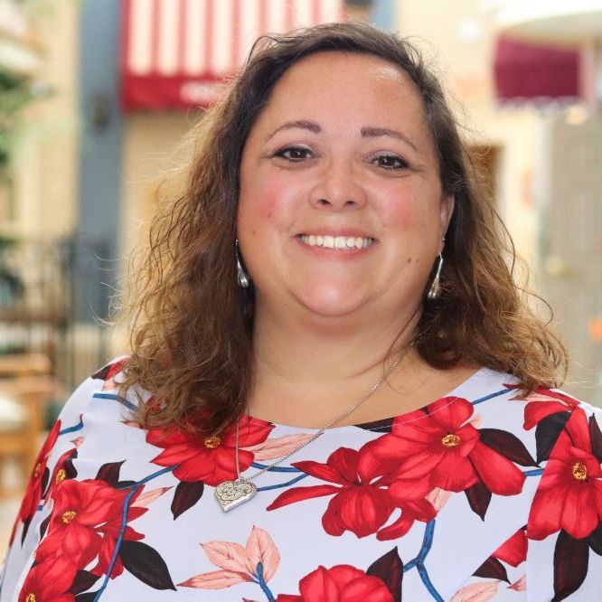 Woman smiling, wearing floral top, standing outdoors in front of a shop.
