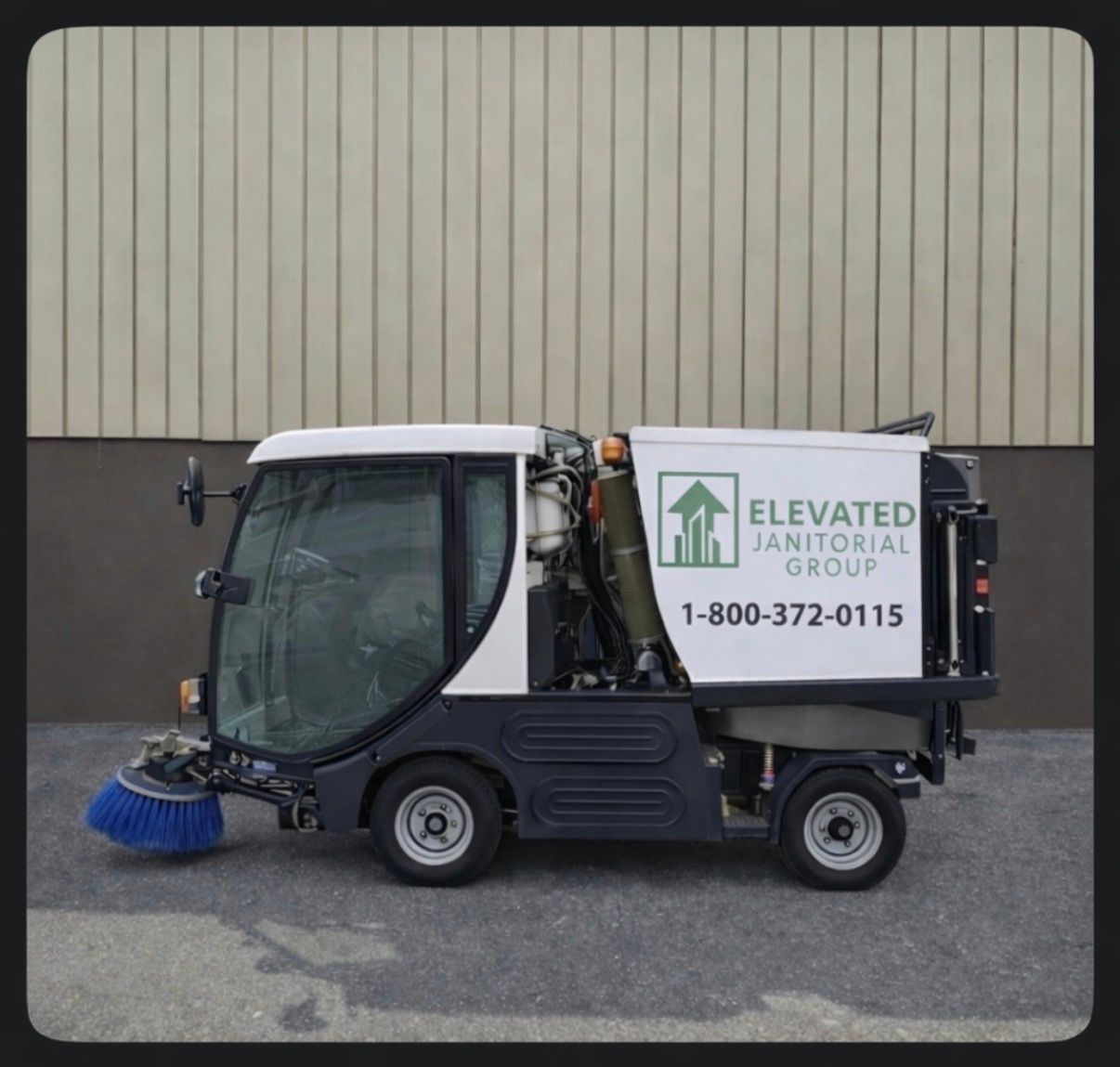 Small black and white street sweeper with blue brush parked beside a beige wall, bearing an Elevated Janitorial sign.