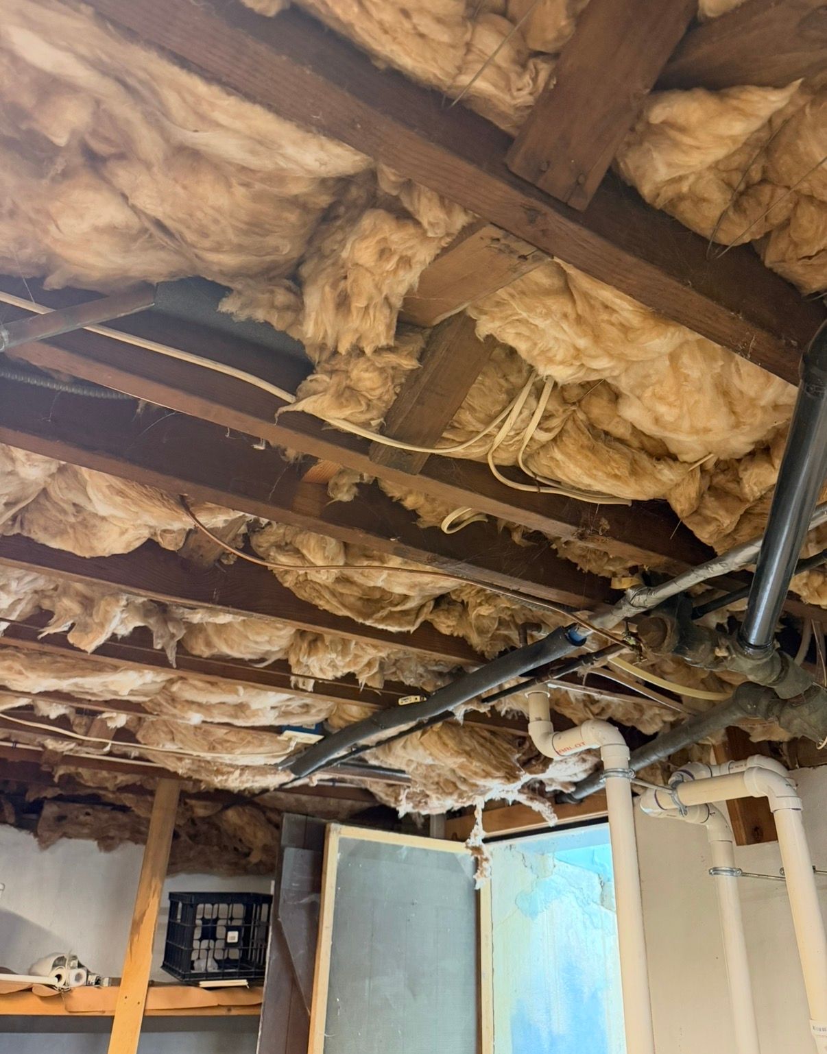 Exposed attic ceiling with insulation, wooden beams, and pipes above an unfinished room.