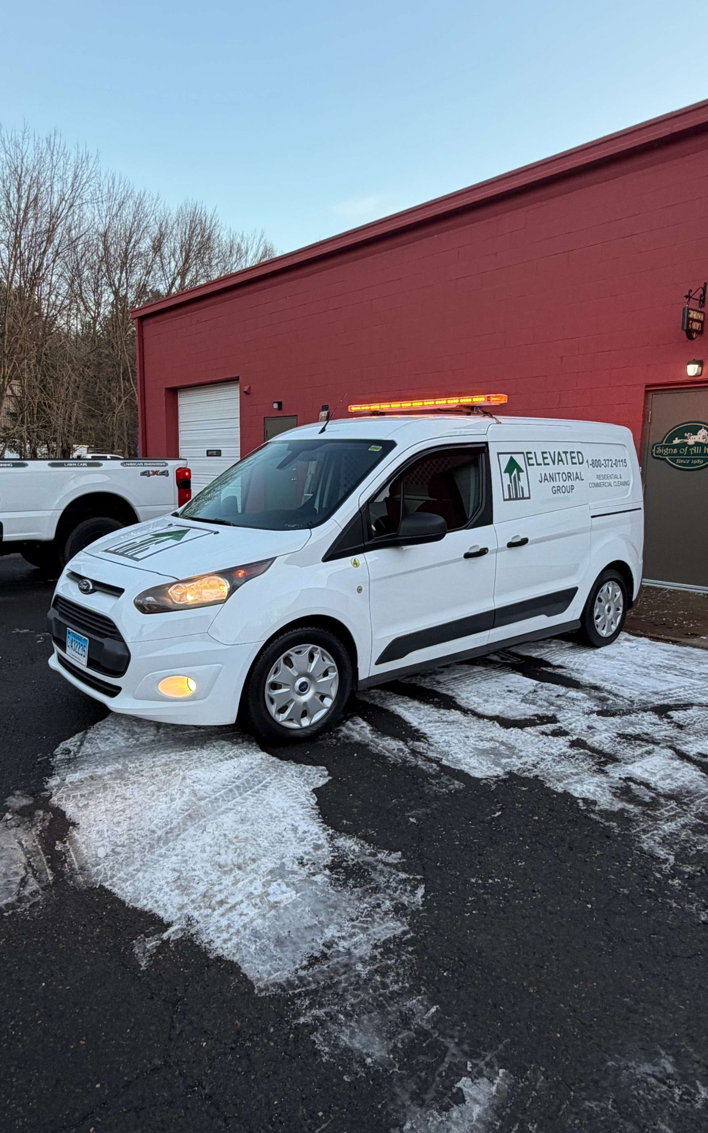 White work van parked near a red building on a snow-covered surface. Amber light bar on top.