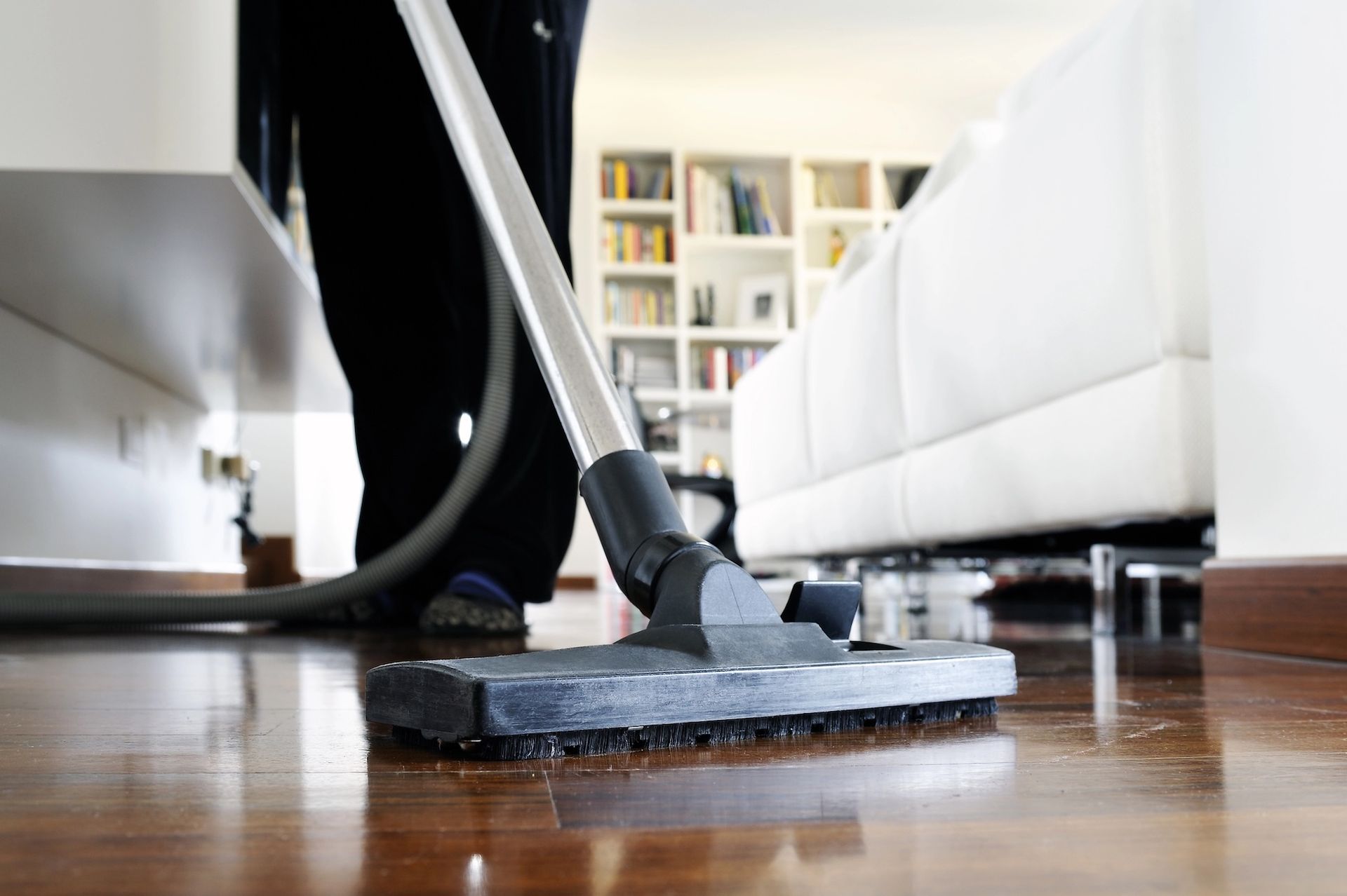 Person vacuuming hardwood floor in a living room with white sofa and bookshelf.