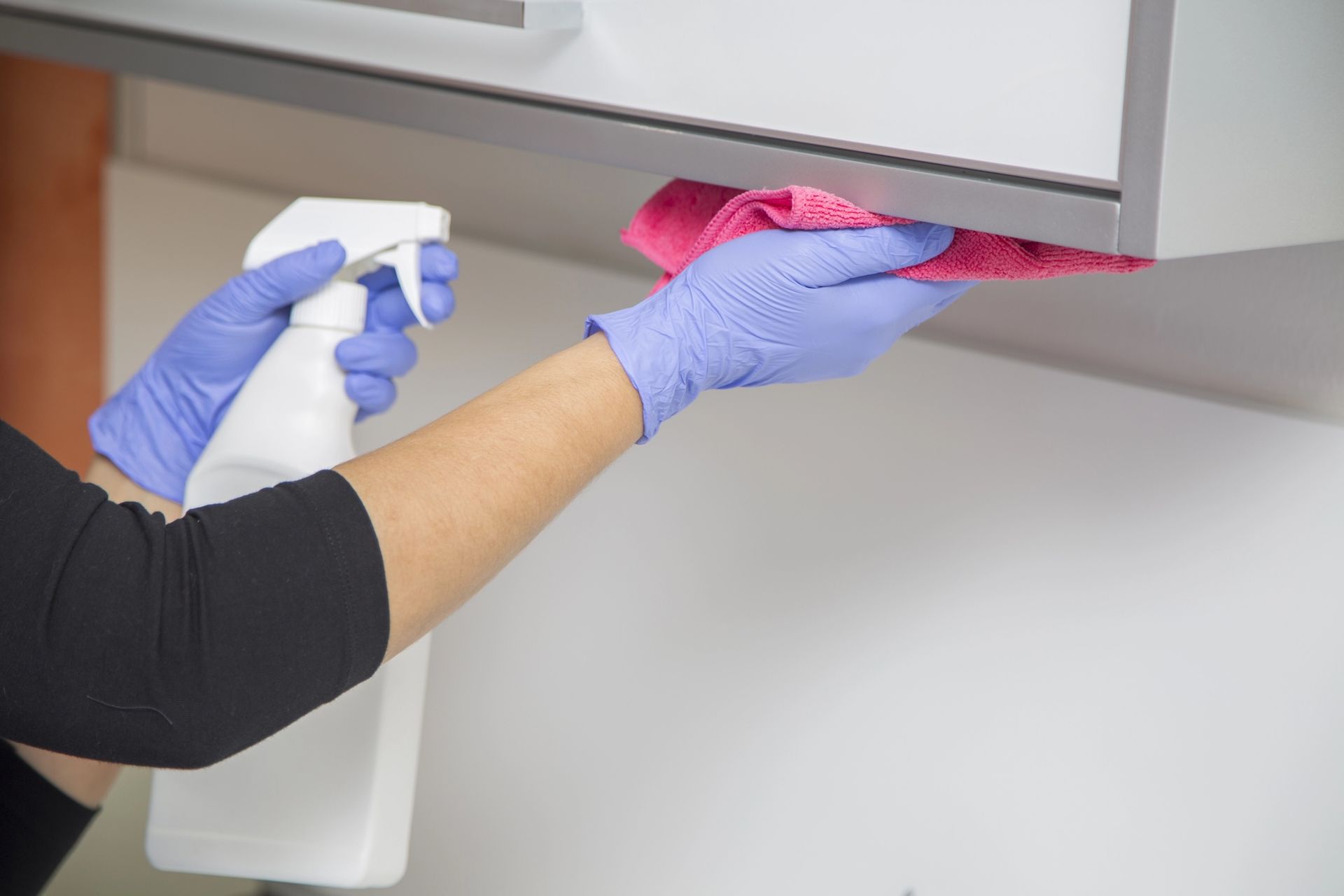 Person wearing gloves cleans underside of kitchen cabinet with spray bottle and cloth.