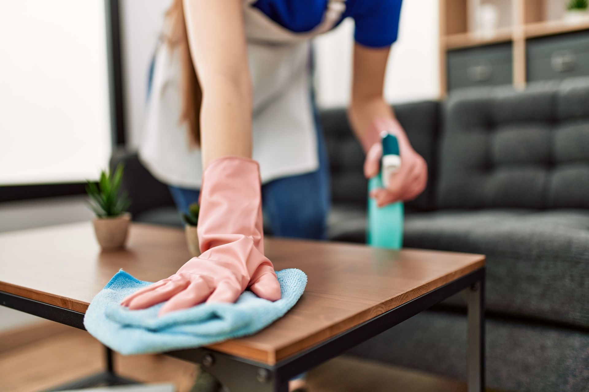 A person wearing a pink glove sprays and wipes a wooden table with a blue cloth in a living room.