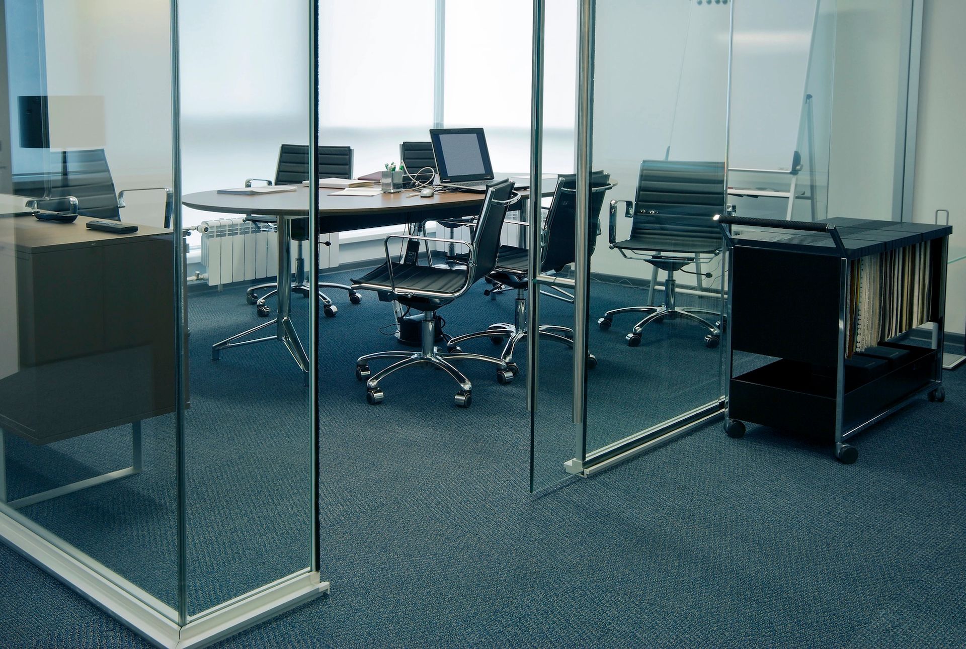 Glass-walled conference room with round table, black chairs, computer monitors, and rolling file cart on blue carpet.