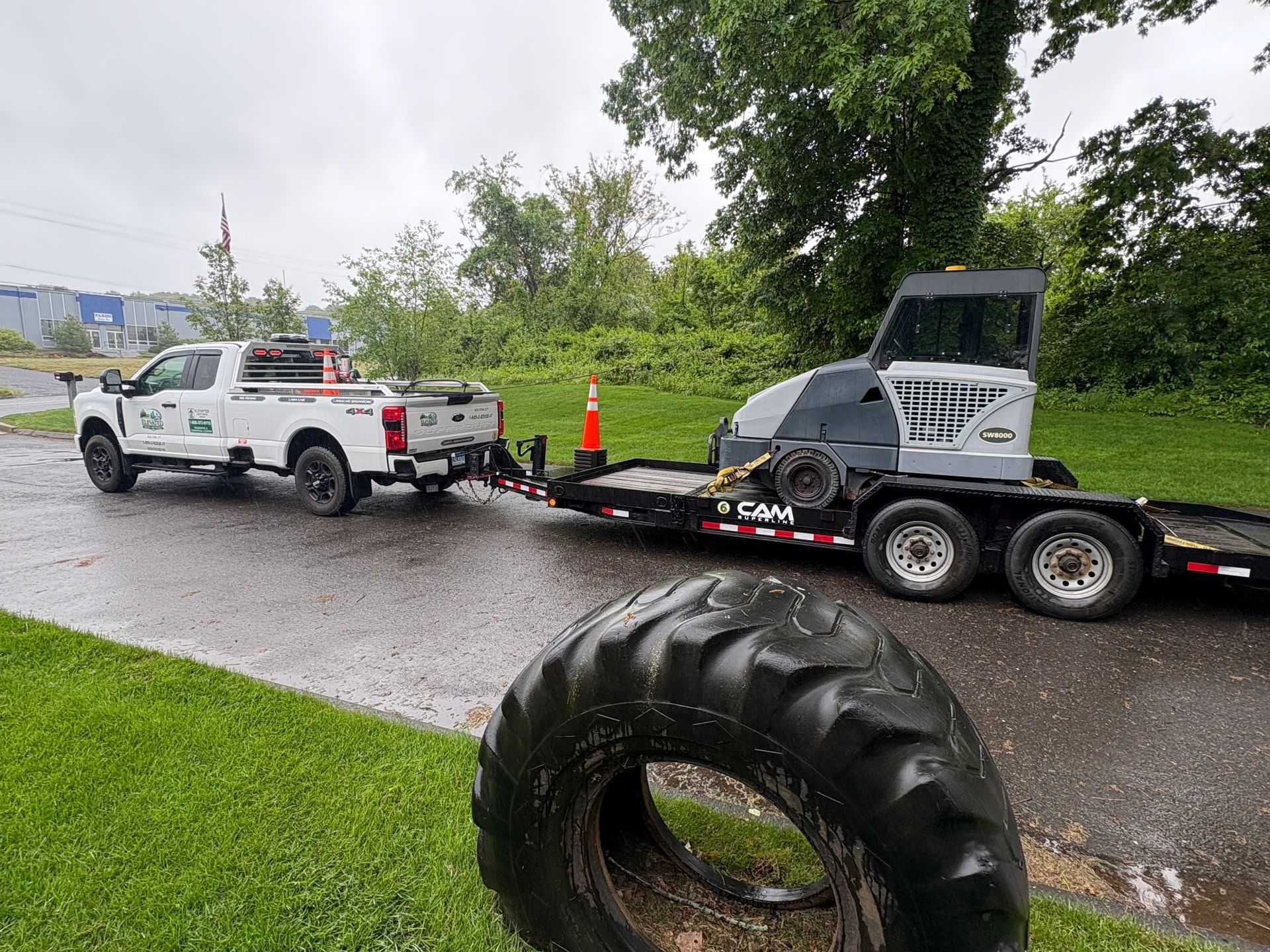 White pickup towing a black skid-steer on a trailer beside a large tire by a wet roadside