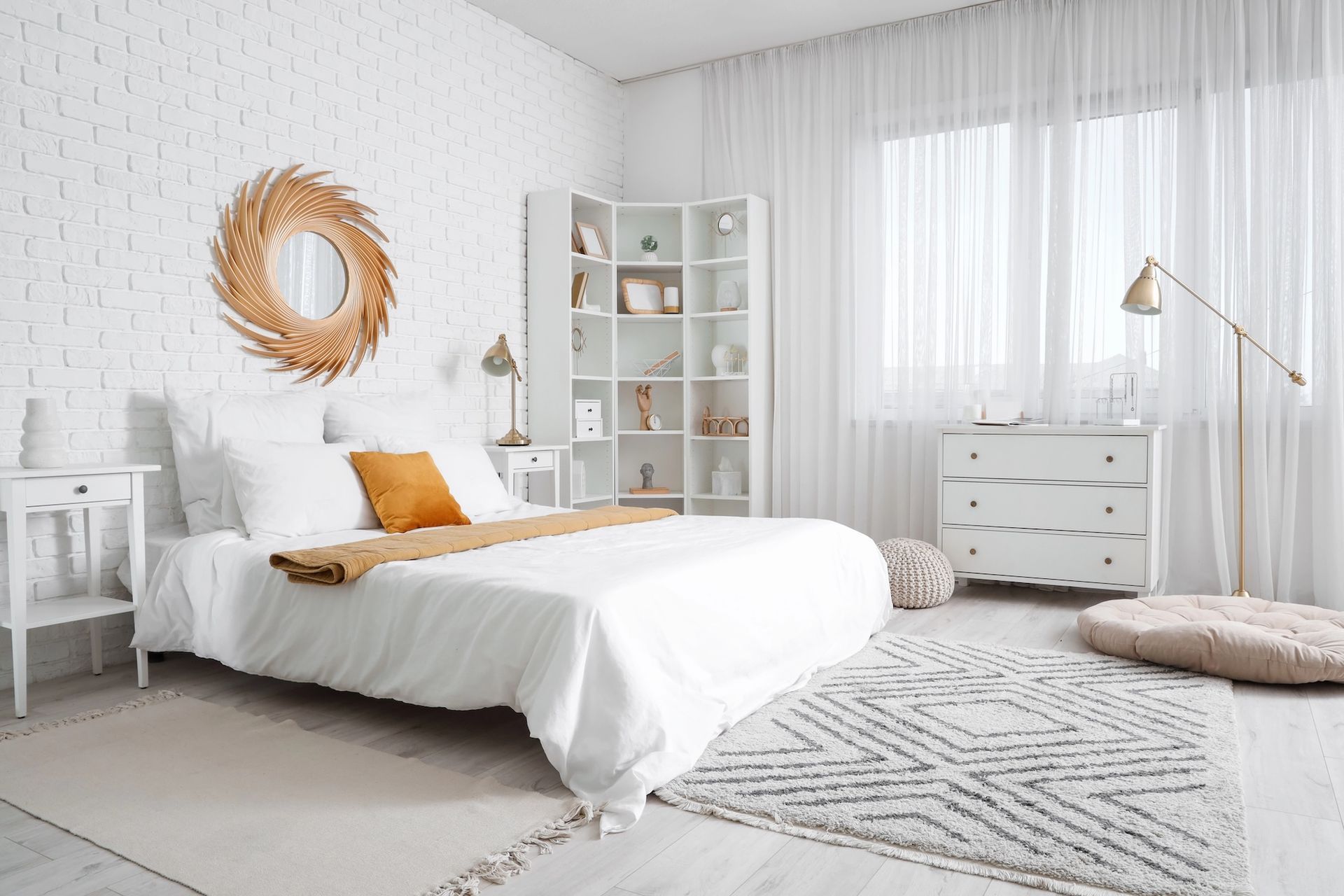 Bright white bedroom with a bed, dresser, shelving, and window. Woven mirror above the bed, a rug, and soft lighting.