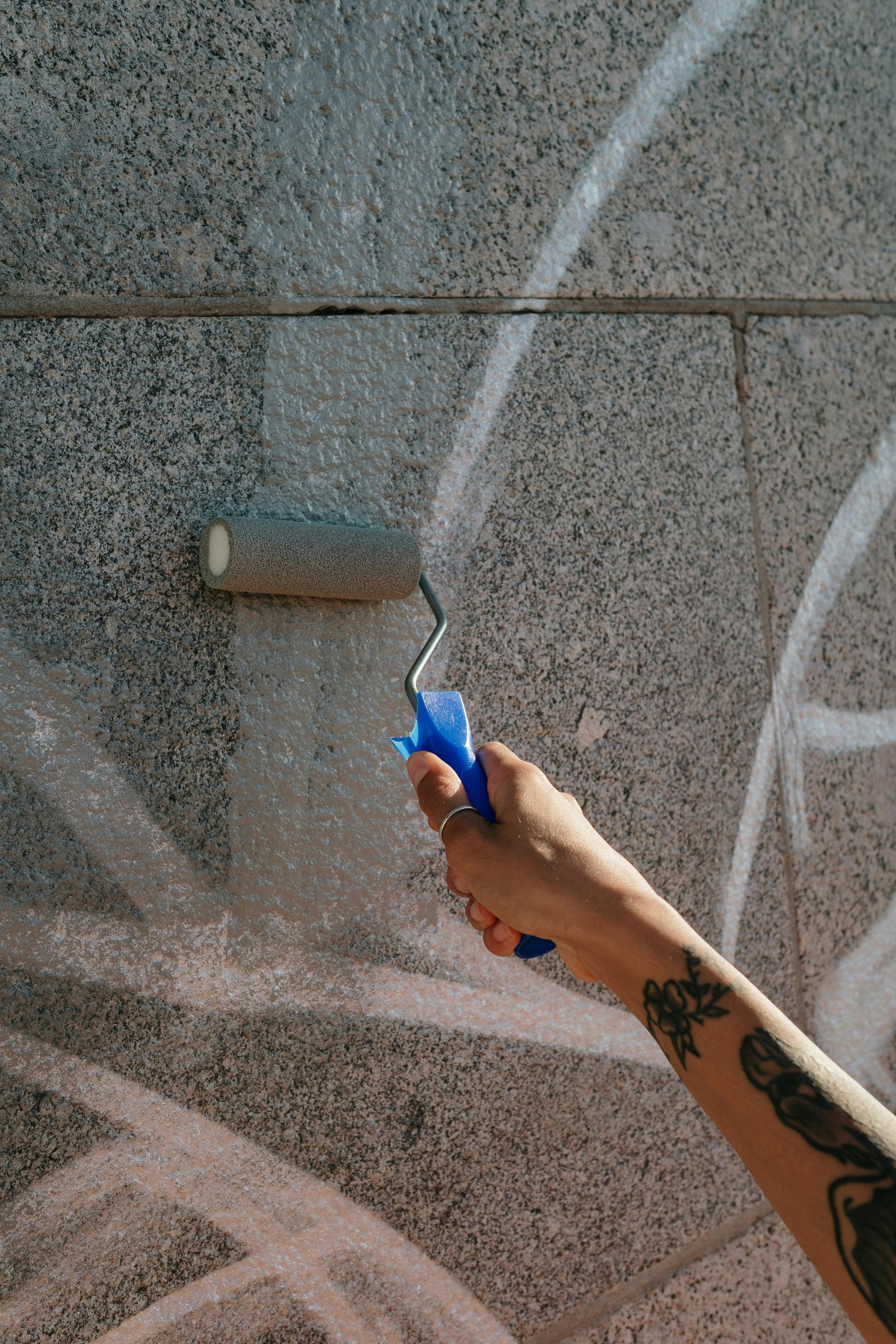 Person painting a gray brick wall with a blue paint roller; arm with tattoos visible.