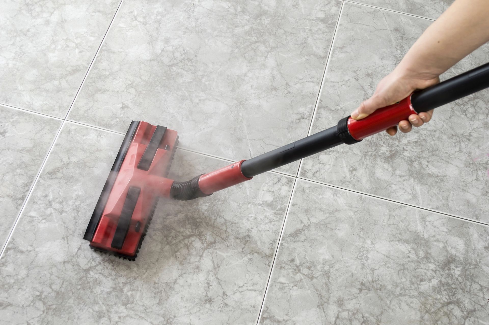 Person using a red and black steam mop on gray tiled floor.
