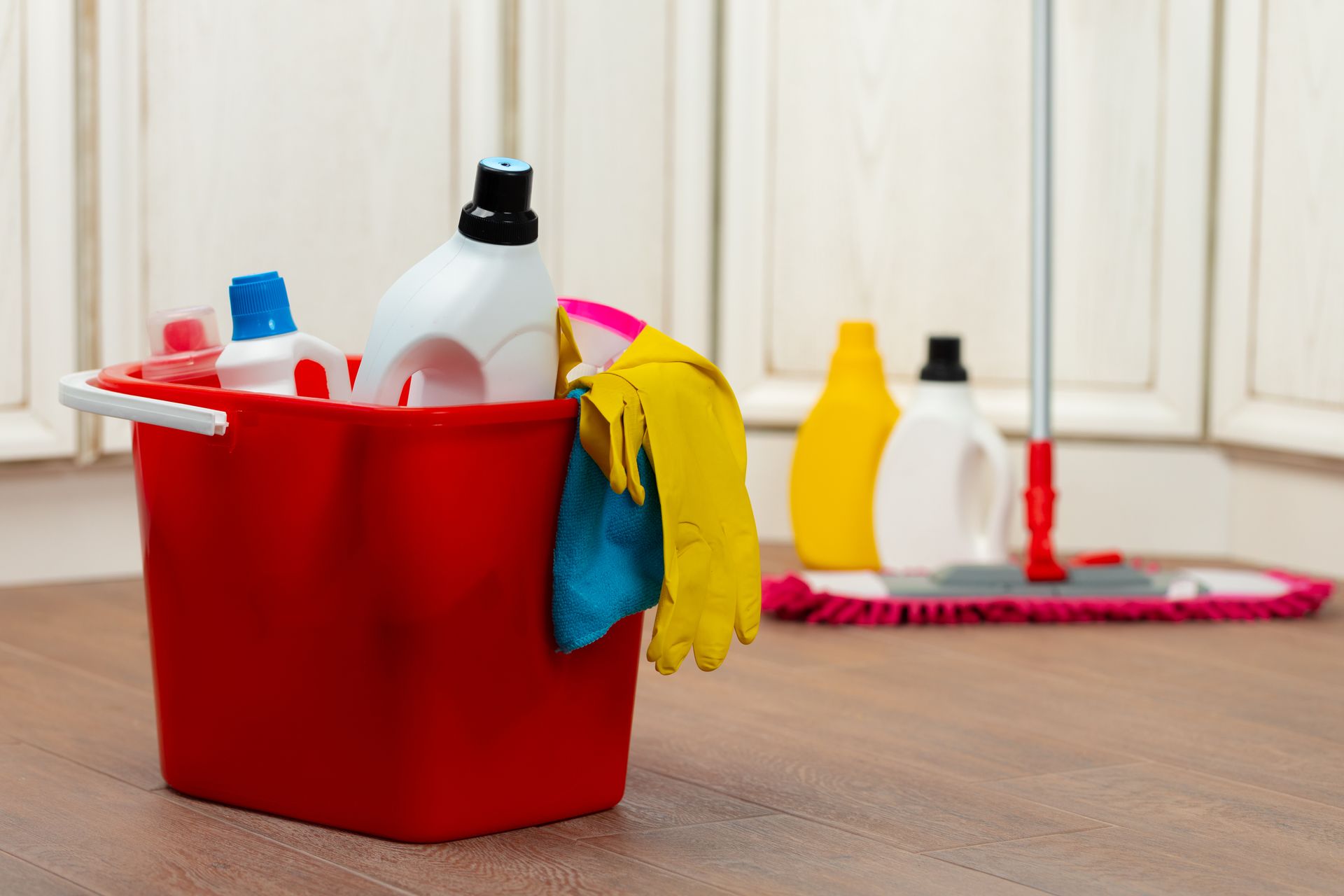 Red cleaning bucket with cleaning supplies on wood floor; mop and bottles in background.