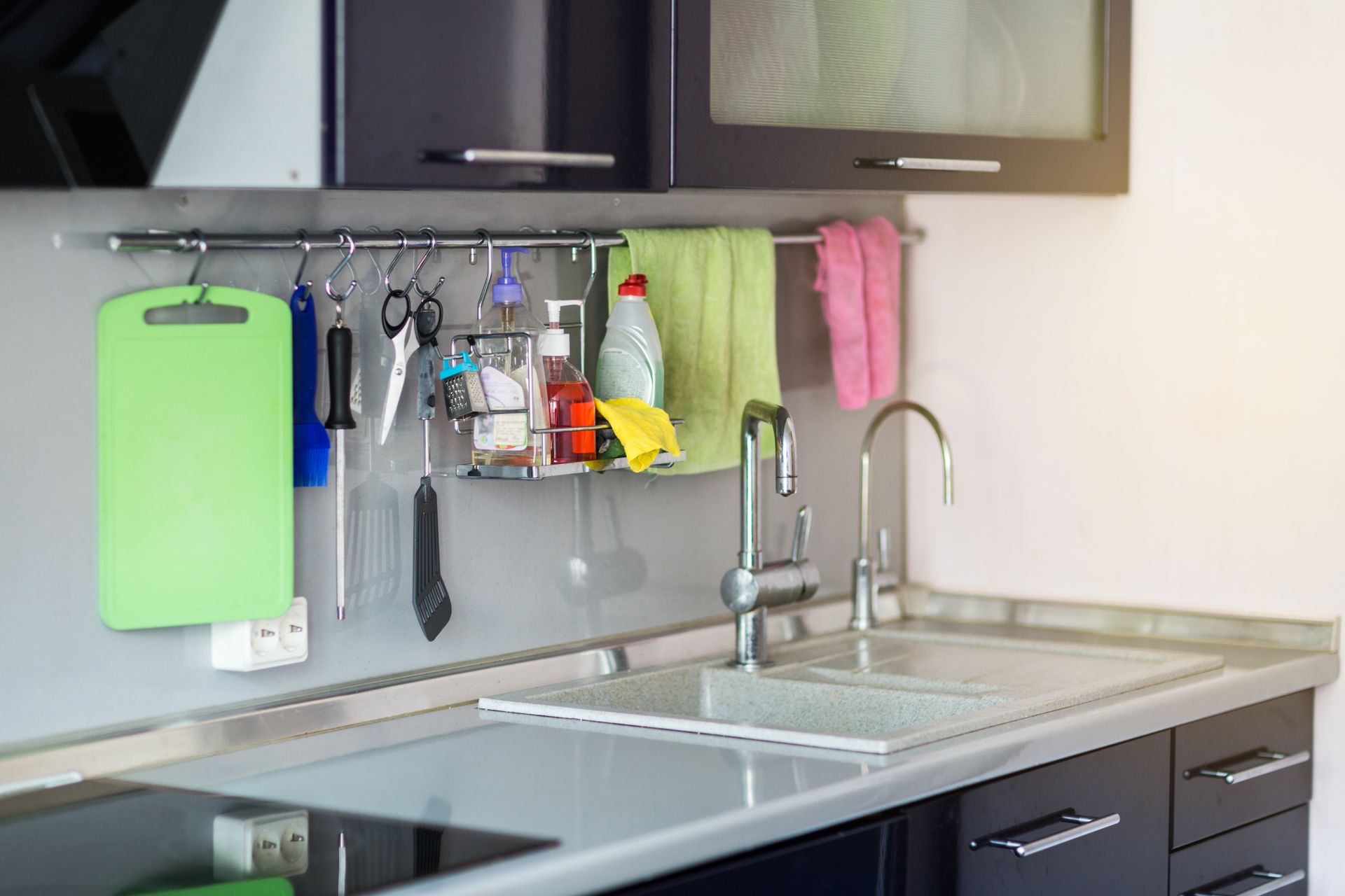 Kitchen counter with sink, hanging rack with cleaning supplies, cutting board, and towels.