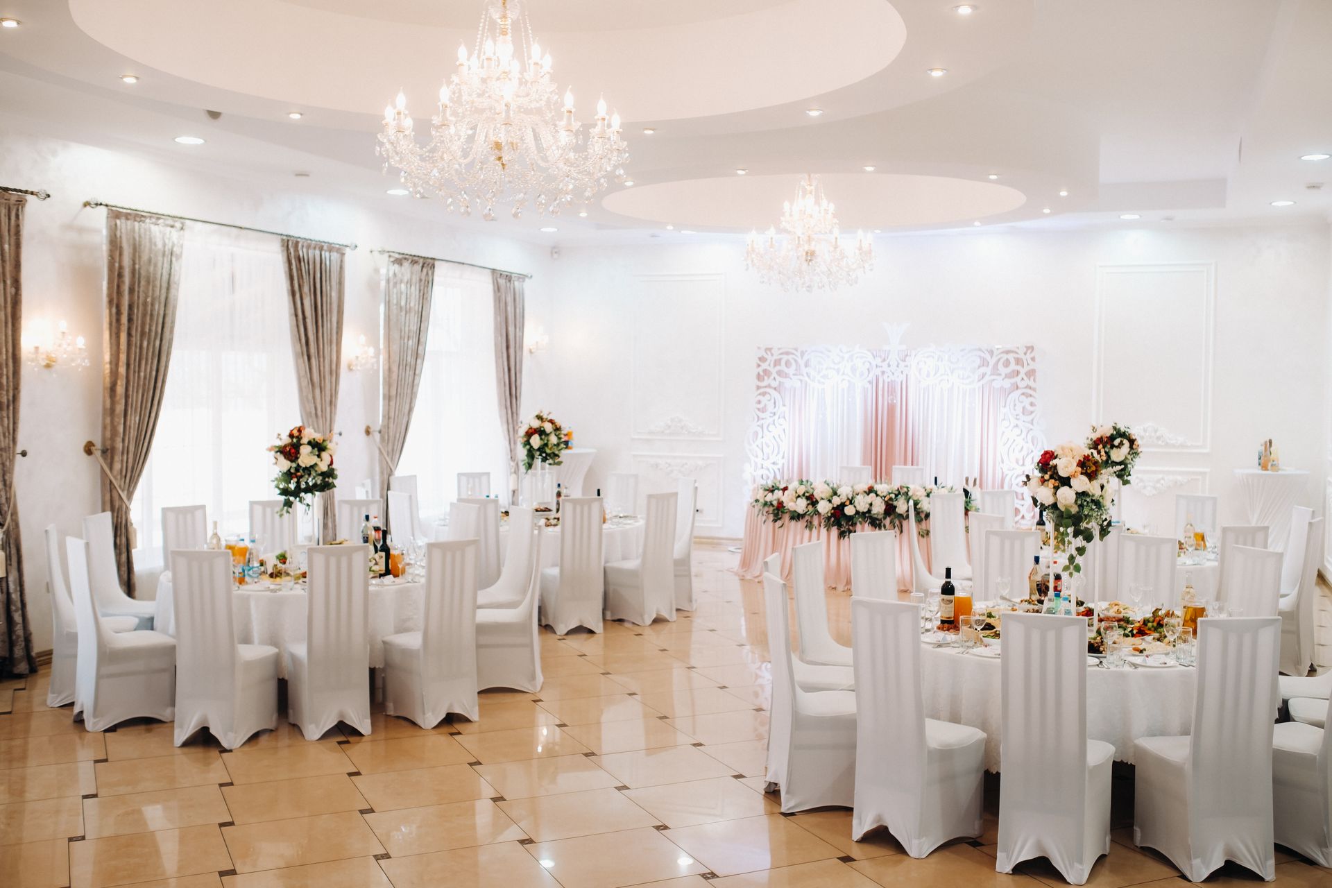 Elegant white ballroom set for a wedding; tables with floral centerpieces, draped curtains, and chandeliers.