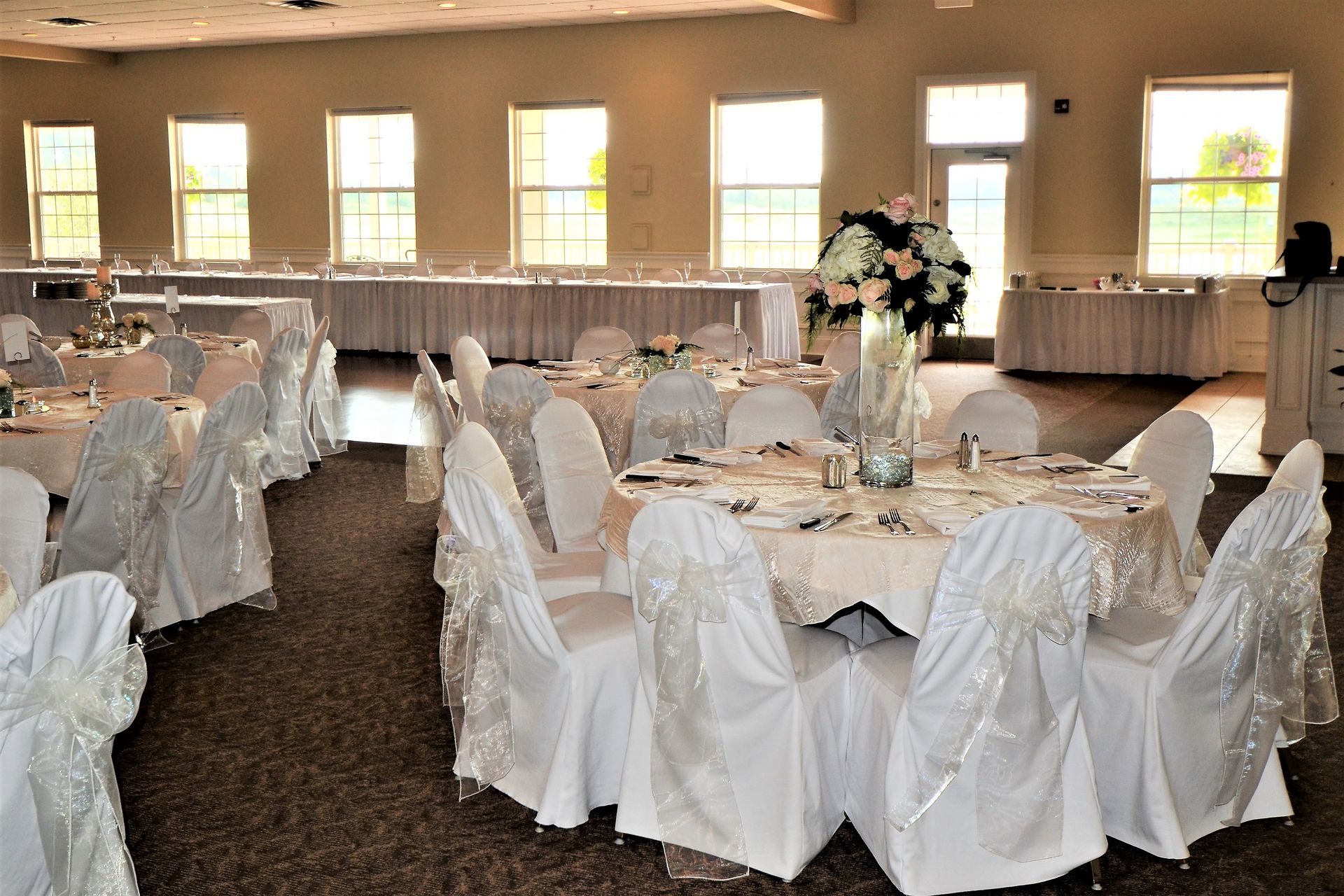 Formal event room with round tables set for a celebration; white chair covers, floral centerpiece, windows.