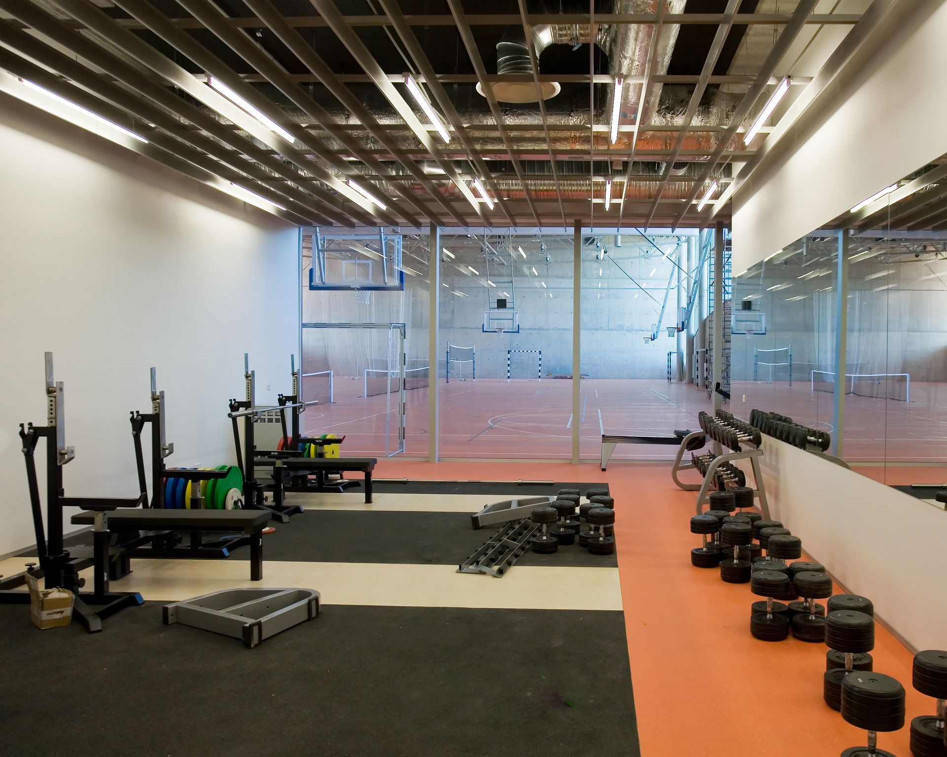 Weight room with barbells, dumbbells, and mirrors, overlooking an empty gymnasium.