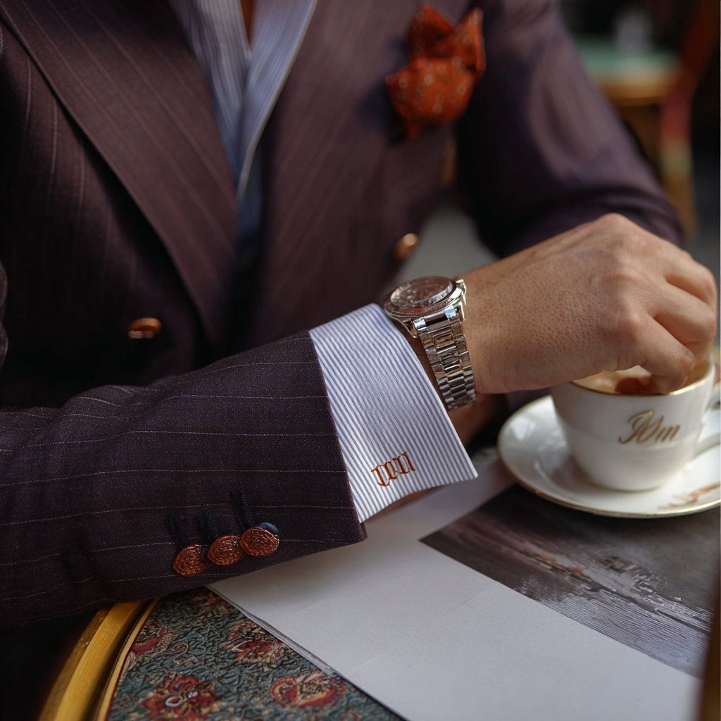 Un hombre con un traje oscuro apoya una muñeca adornada con un reloj junto a una taza y un platillo en una mesa de café.