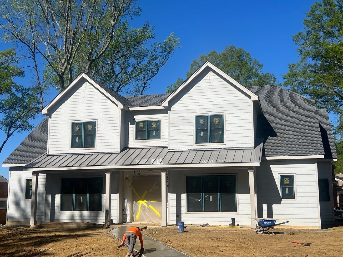 White two-story house with black shutters and a front porch under construction, surrounded by trees and blue sky
