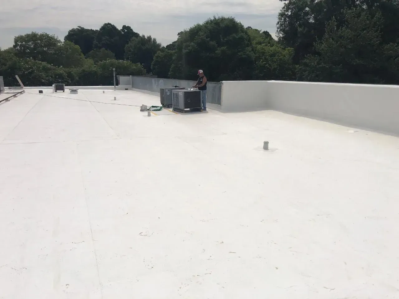 Flat white rooftop with a worker near HVAC units and trees in the background.