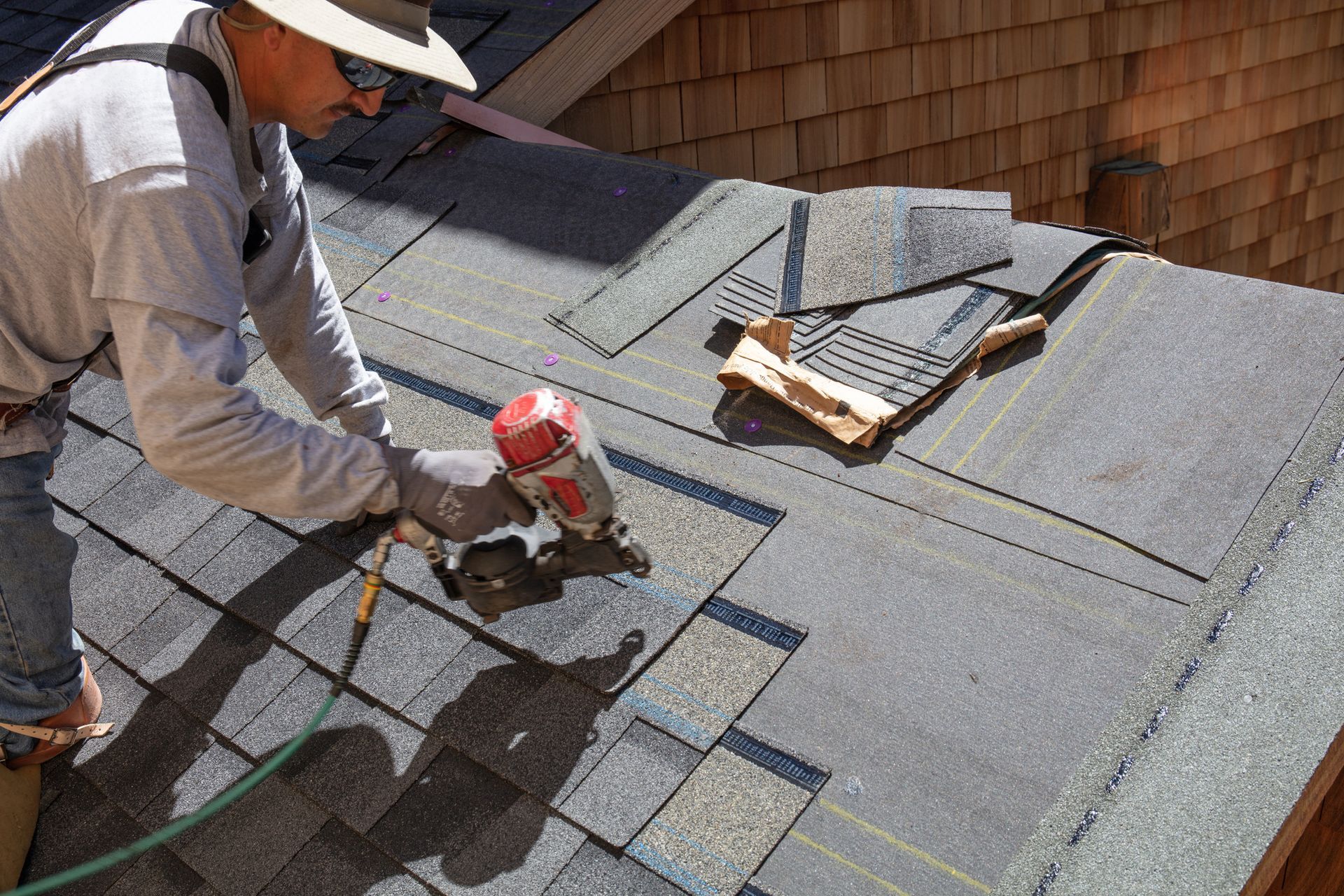 Worker using a nail gun to install shingles on a roof, with tools nearby
