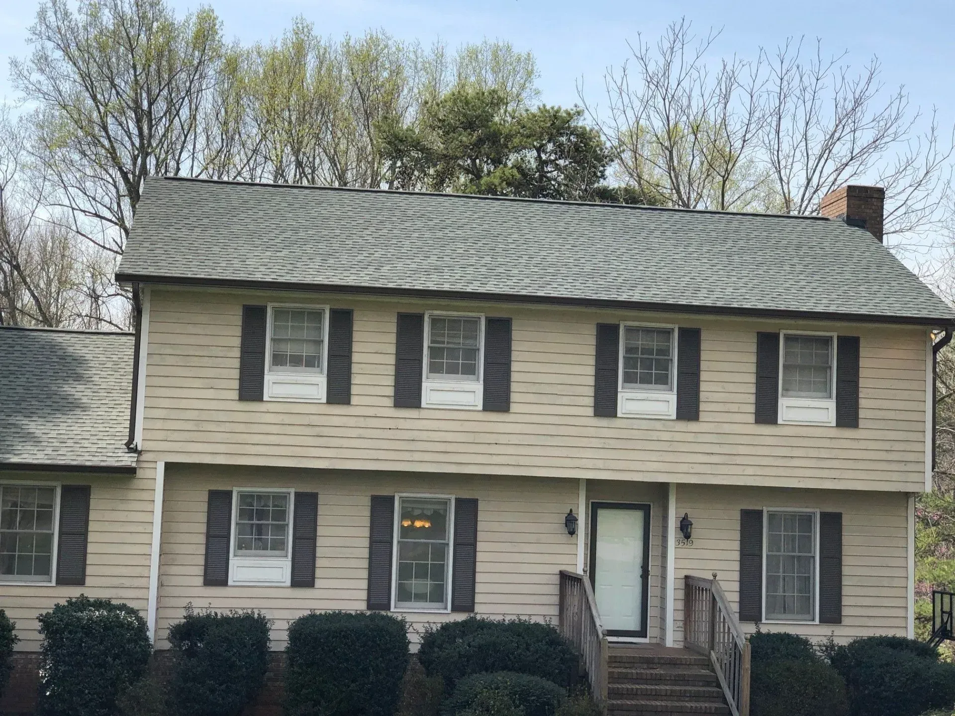 Two-story beige house with dark shutters, a centered front door, and leafless trees behind it.