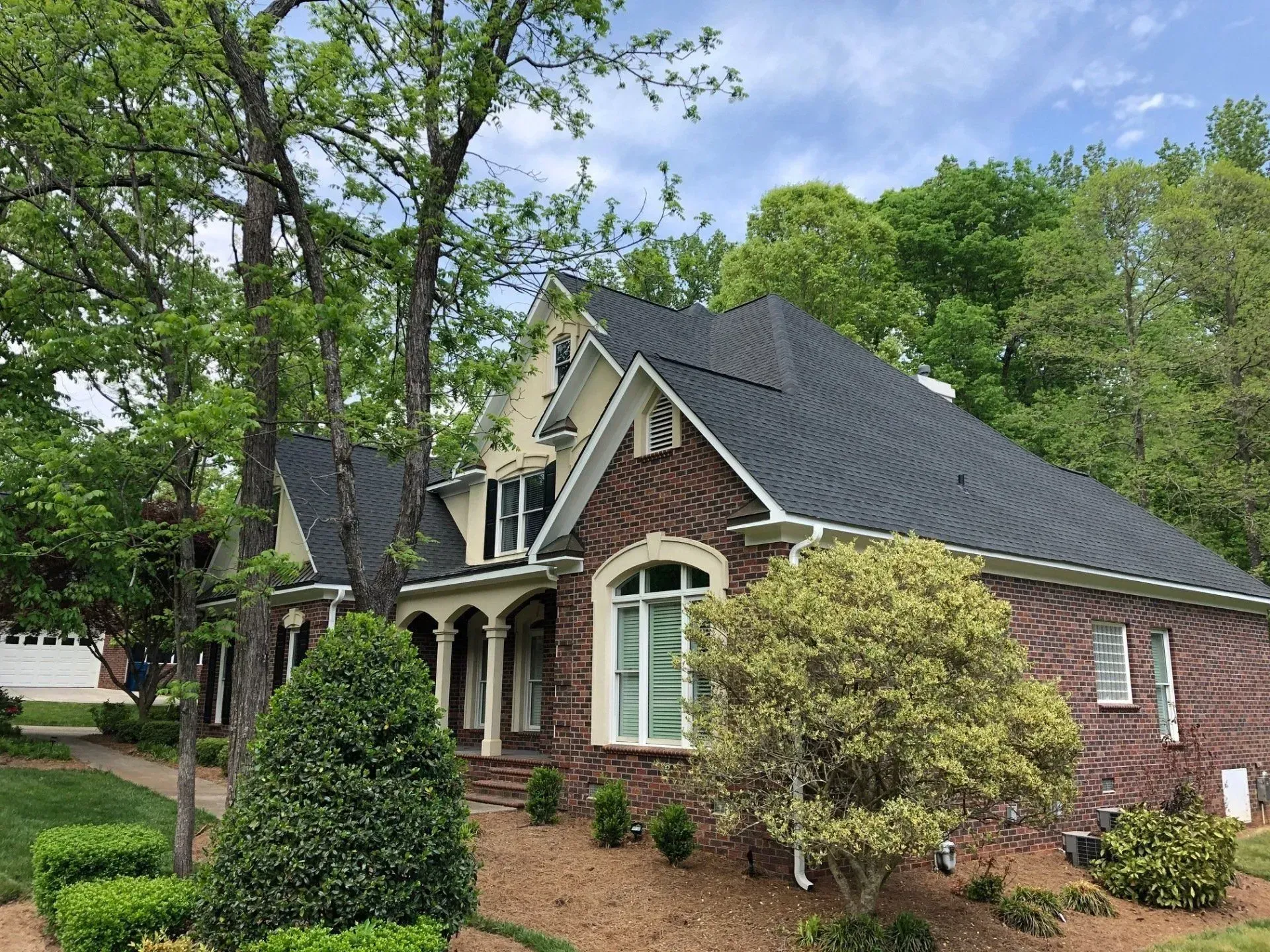 Brick house with dark gray roof and arched windows, surrounded by trees and shrubs on a sunny day