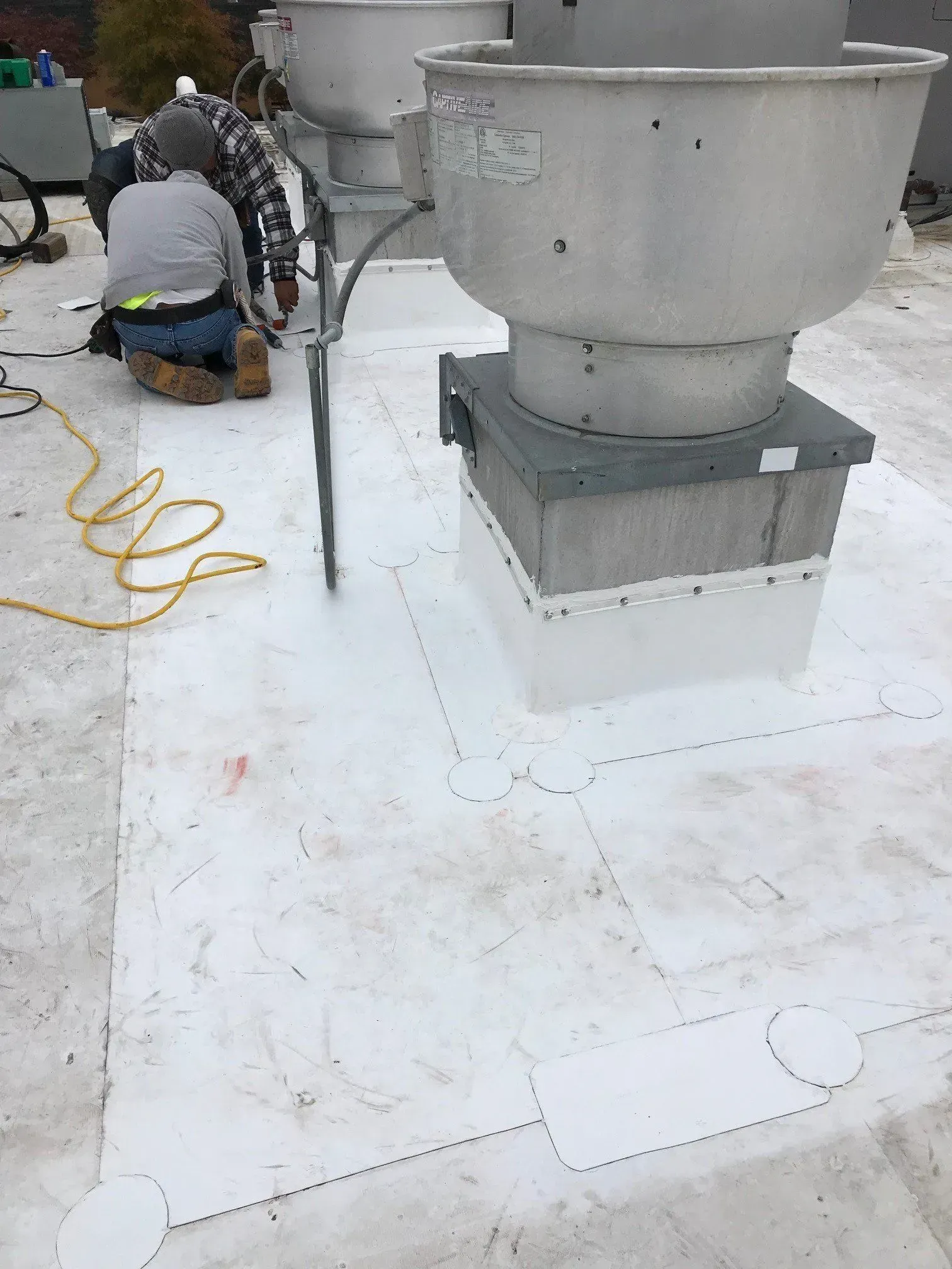 Workers installing equipment on a white rooftop beside a large metal vent and yellow cord.