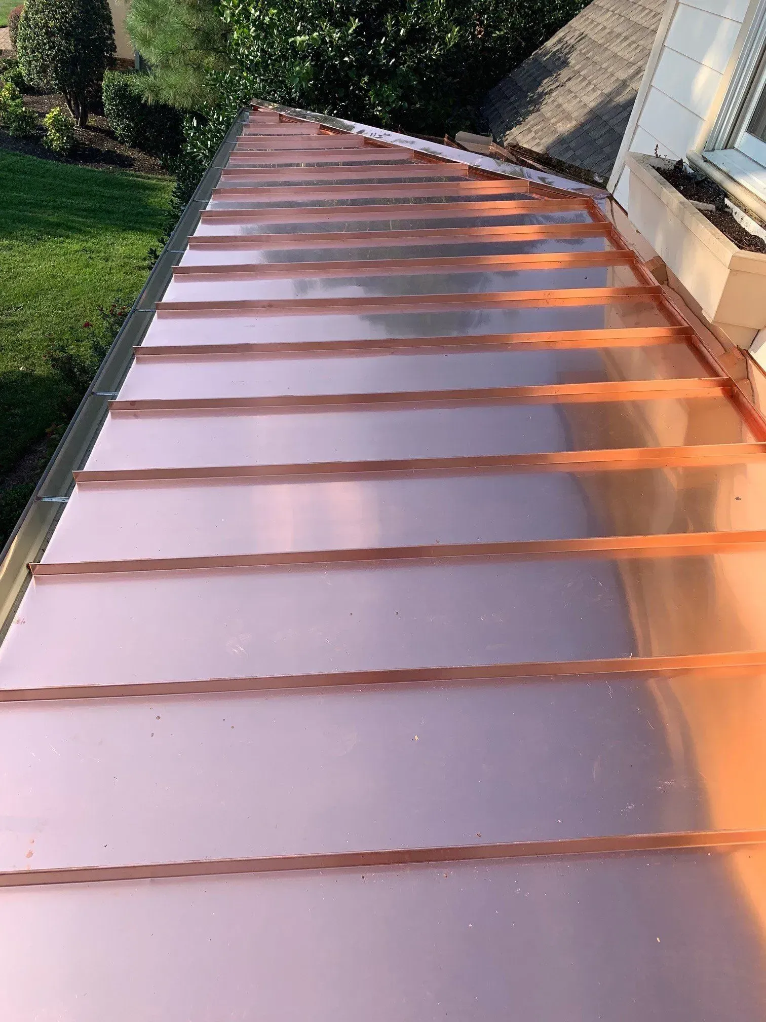 Copper-colored metal roof viewed from above beside a house and greenery