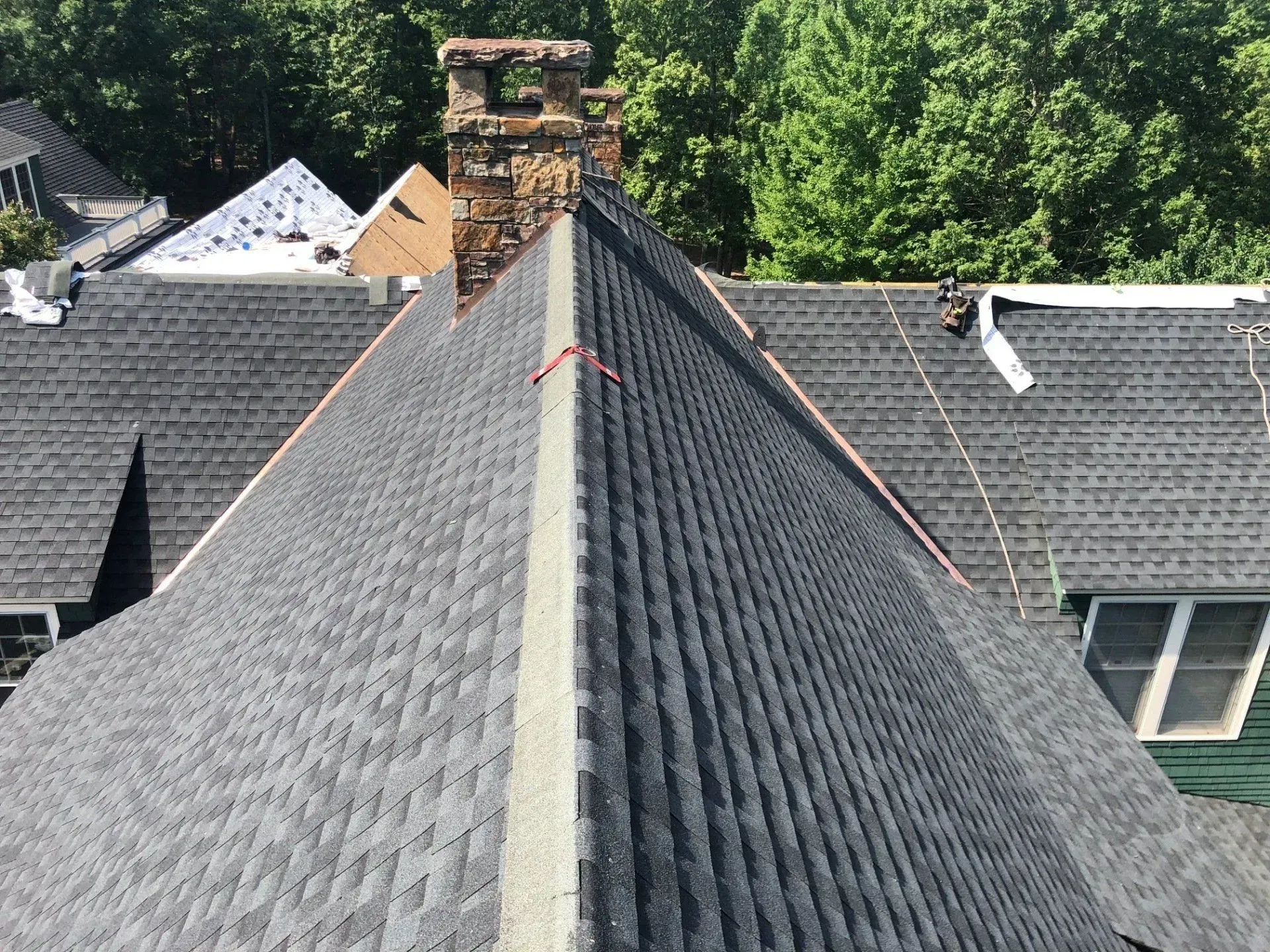 Rooftop ridge with gray shingles and a brick chimney, surrounded by trees and neighboring houses.