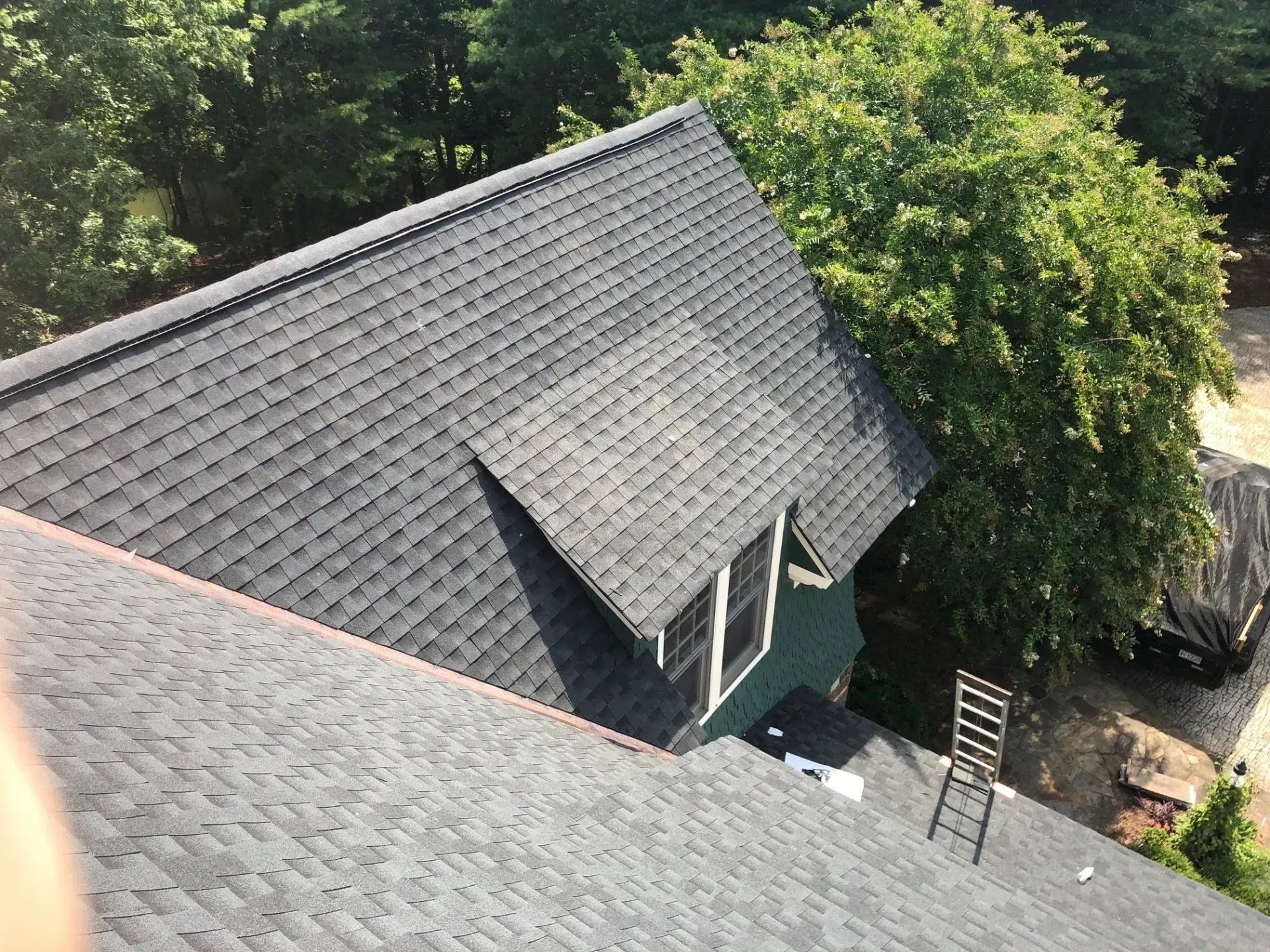 Aerial view of a gray-roofed house surrounded by trees and a driveway with a ladder nearby