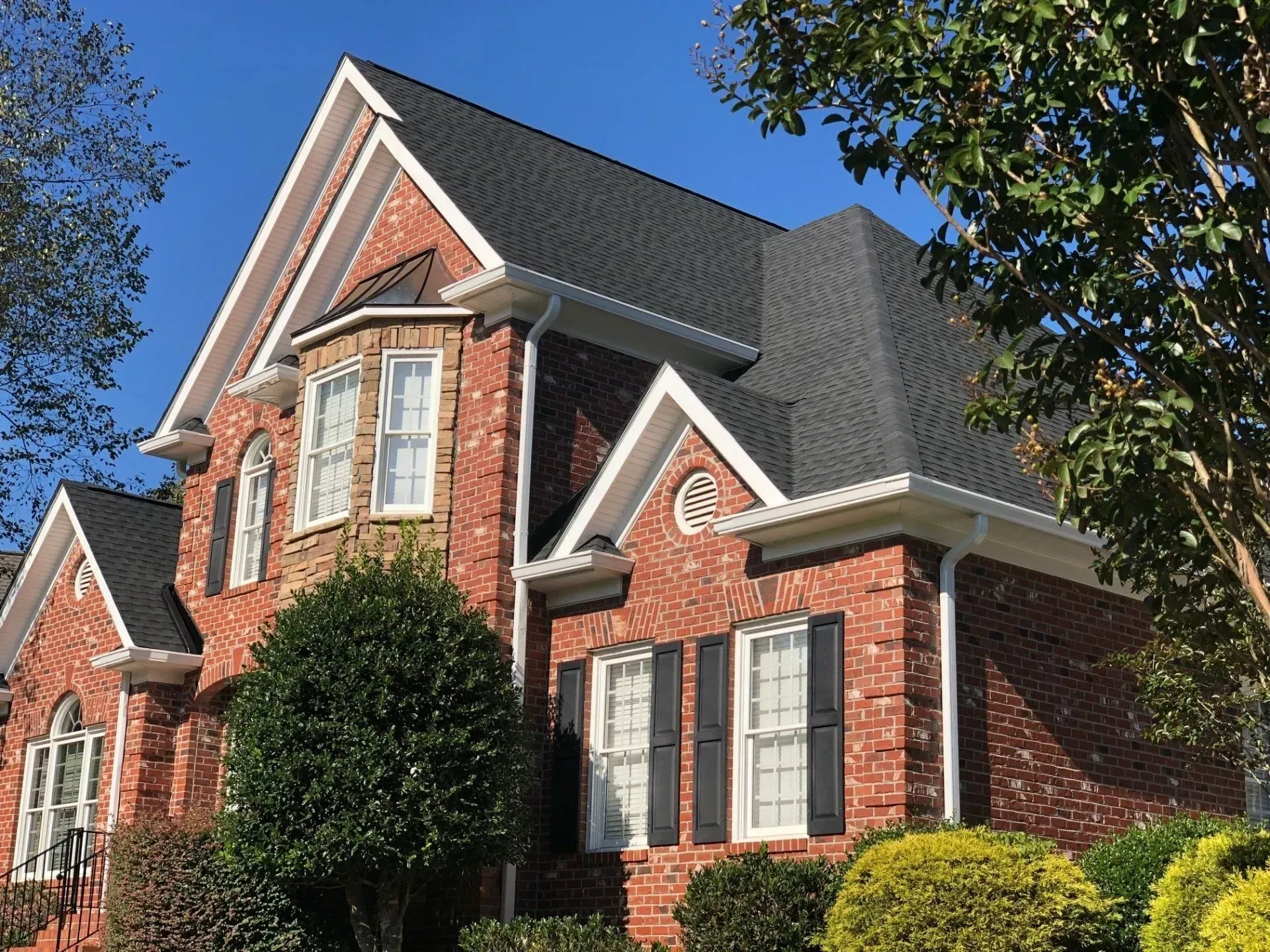 Red brick suburban house with white trim, dark roof, and a large front tree under a blue sky