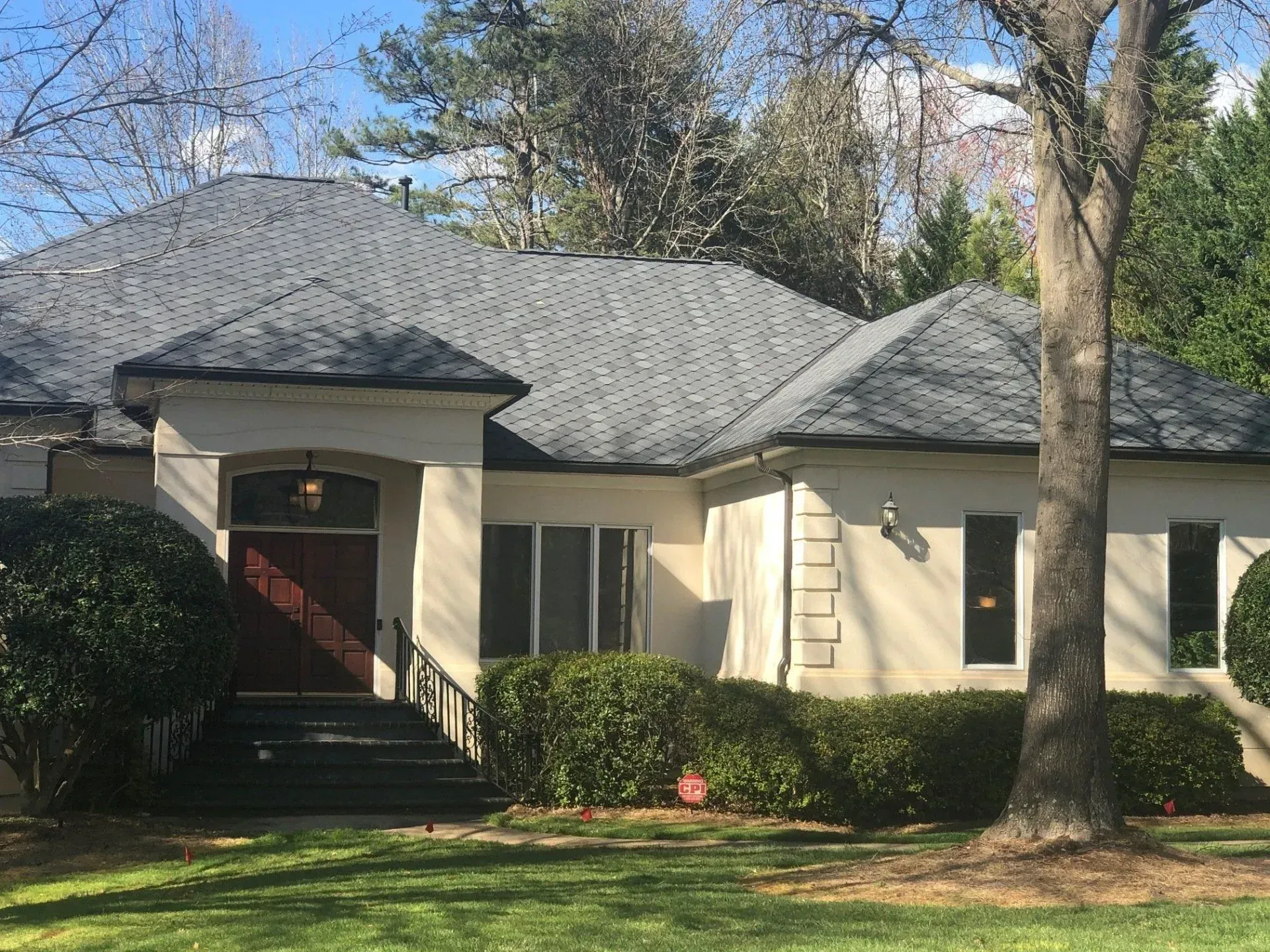 Single-story house with gray roof, cream siding, red front door, and large trees in front