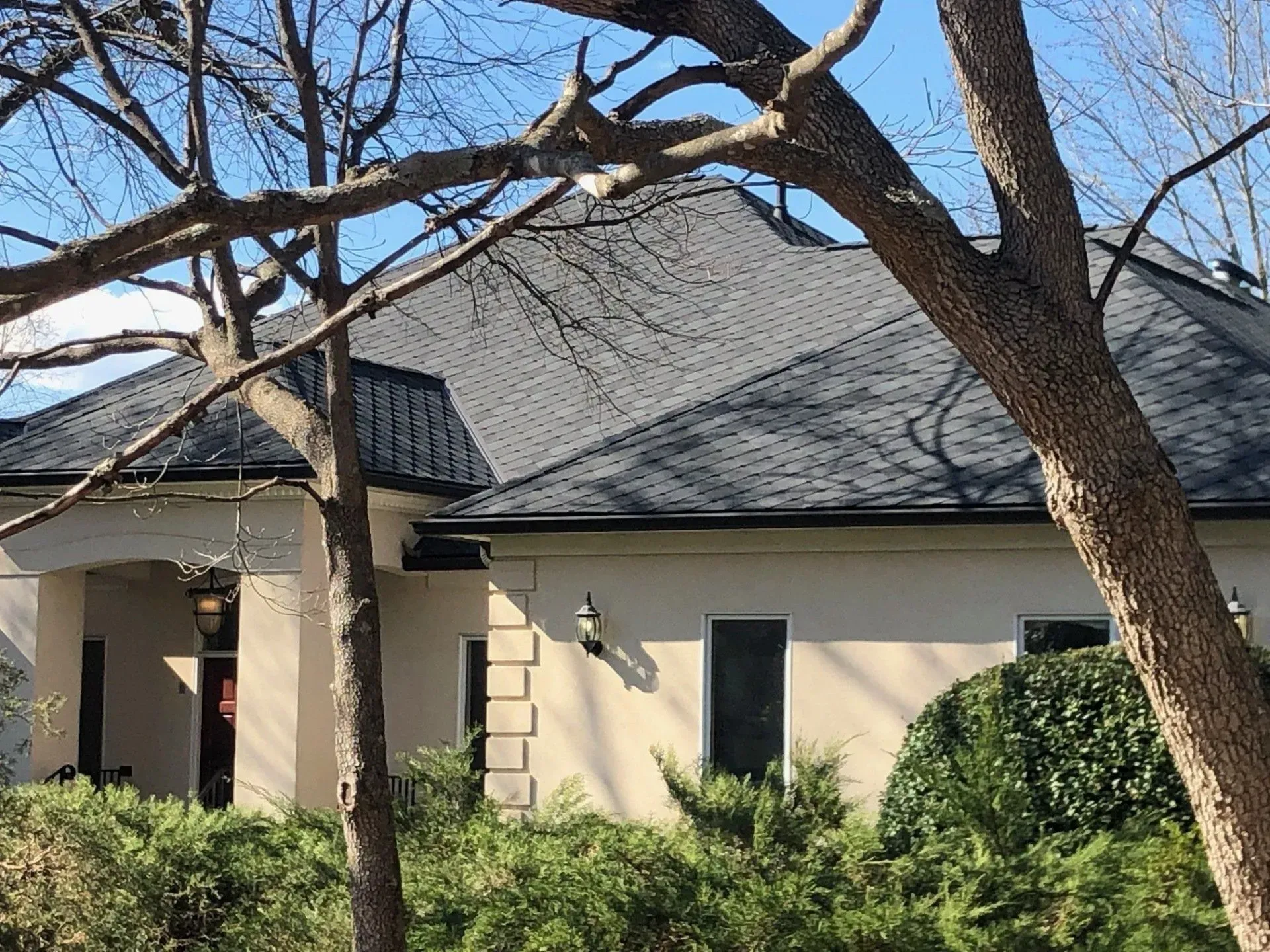 Front of a beige house with a dark roof, framed by leafless trees and green shrubs under a clear blue sky