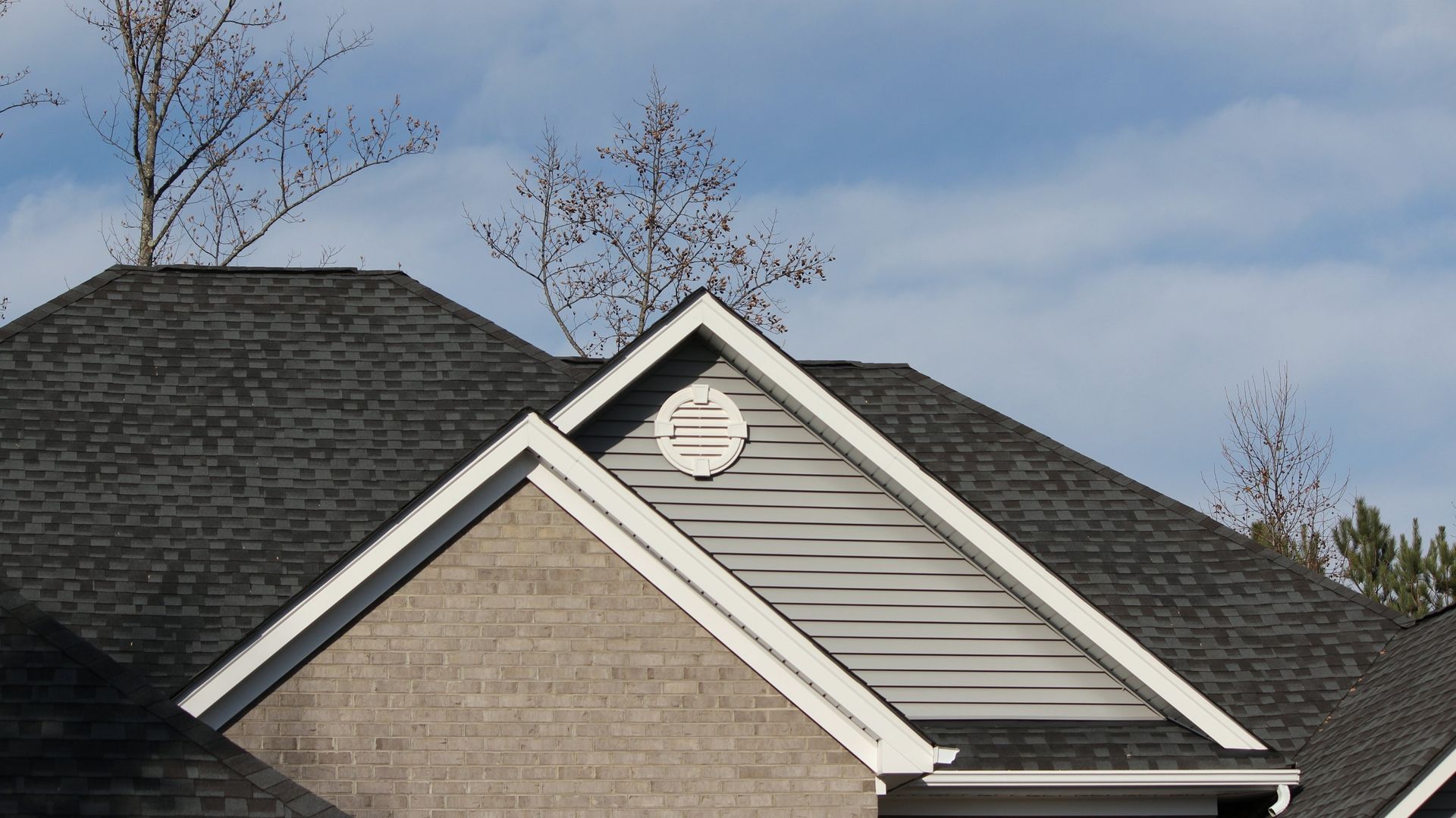 House roof with gray shingles and beige siding under a cloudy sky, with bare trees in the background