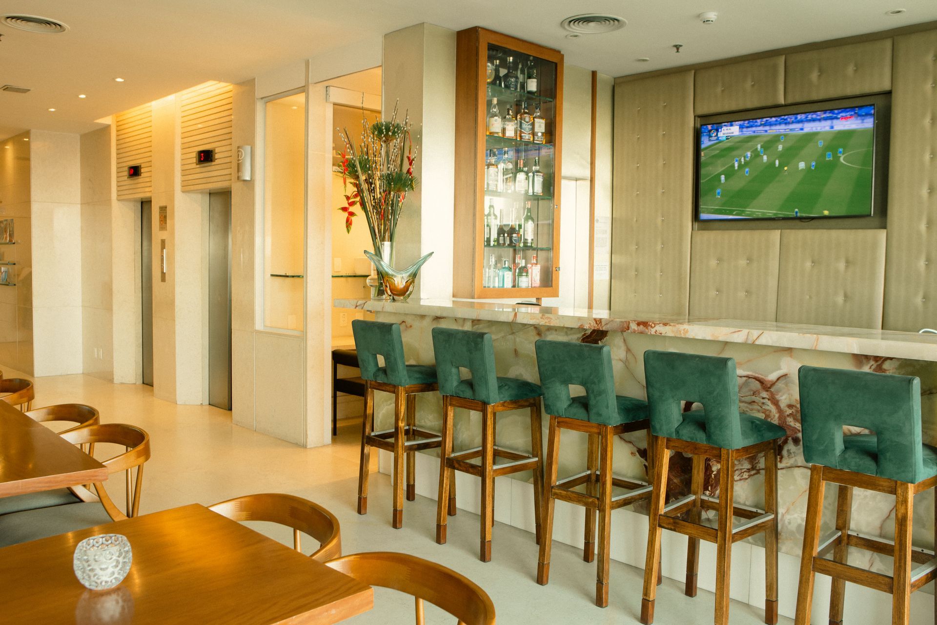 Área de bar com bancos, TV transmitindo um jogo de futebol e mesas; cores neutras com detalhes em verde.