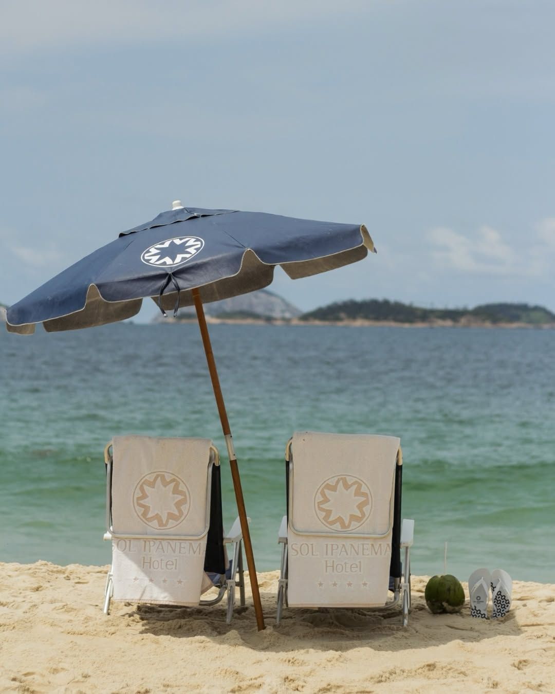 Duas cadeiras de praia sob um guarda-sol azul em uma praia de areia com vista para o oceano.