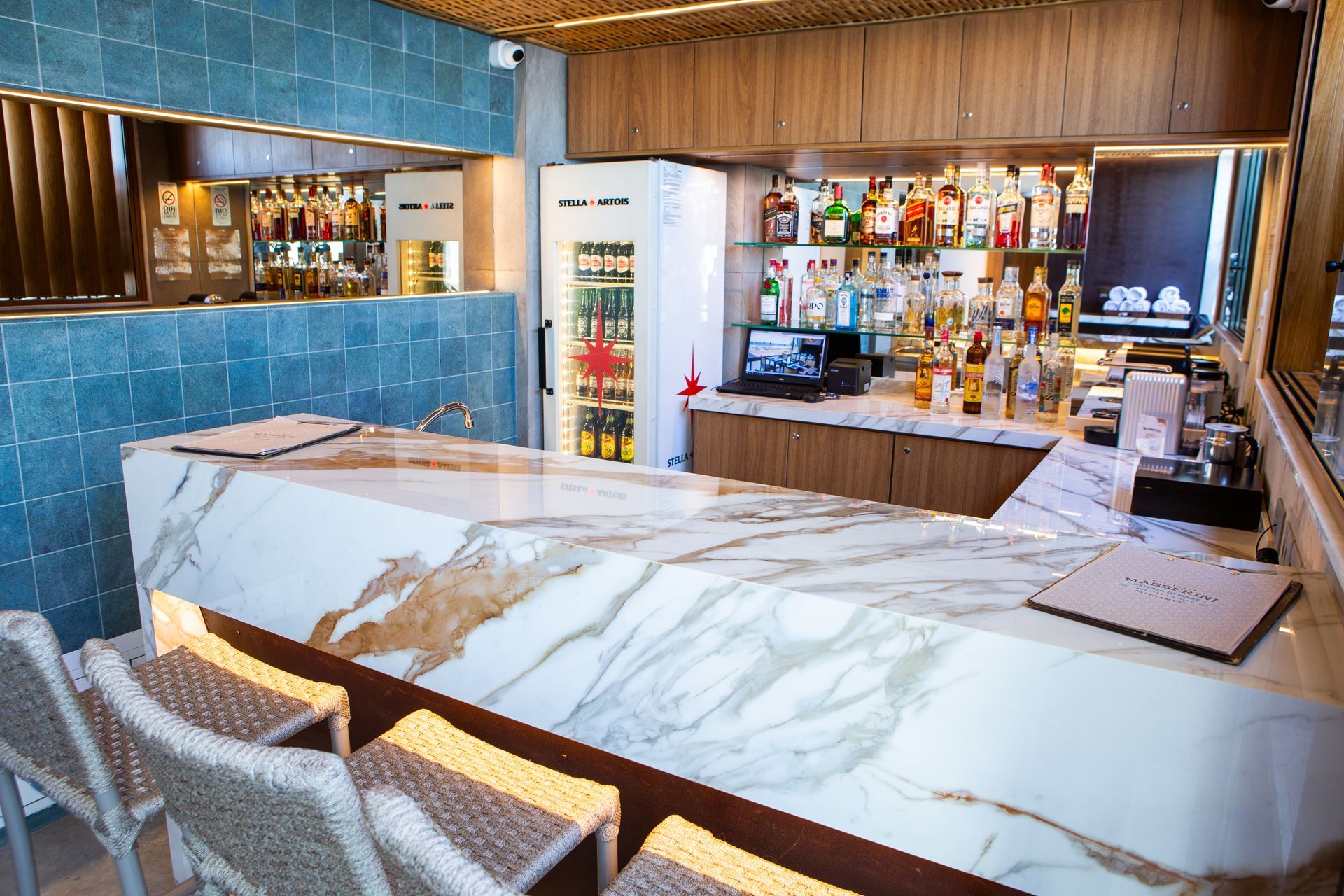 Bar com bancada de mármore, garrafas de bebidas e bancos de vime. Parede com azulejos azuis ao fundo.