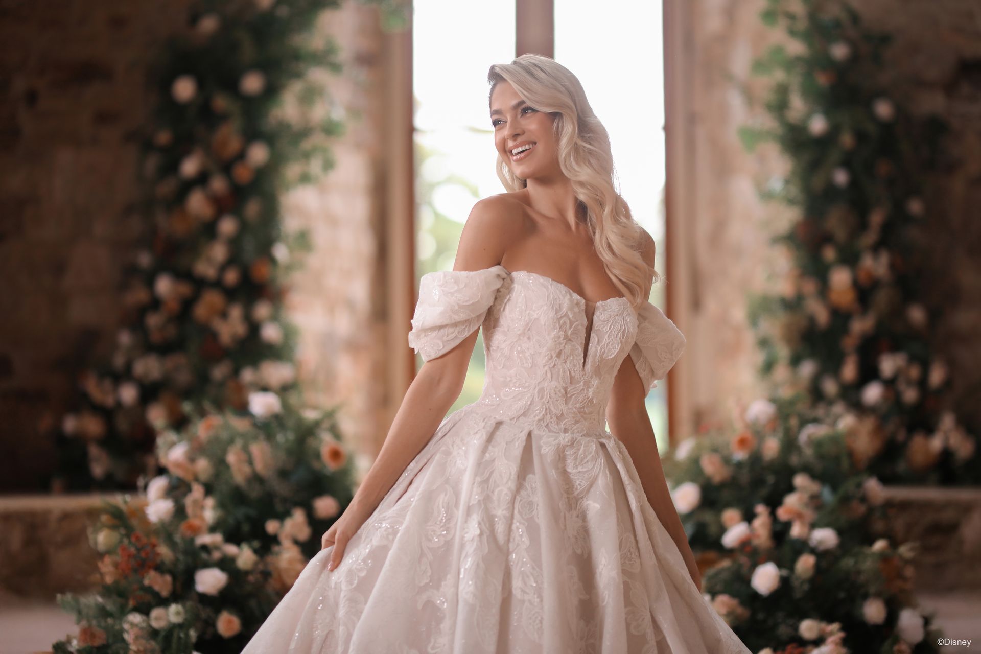Woman in a white lace wedding dress with a large bow and train, standing on a path in a garden.