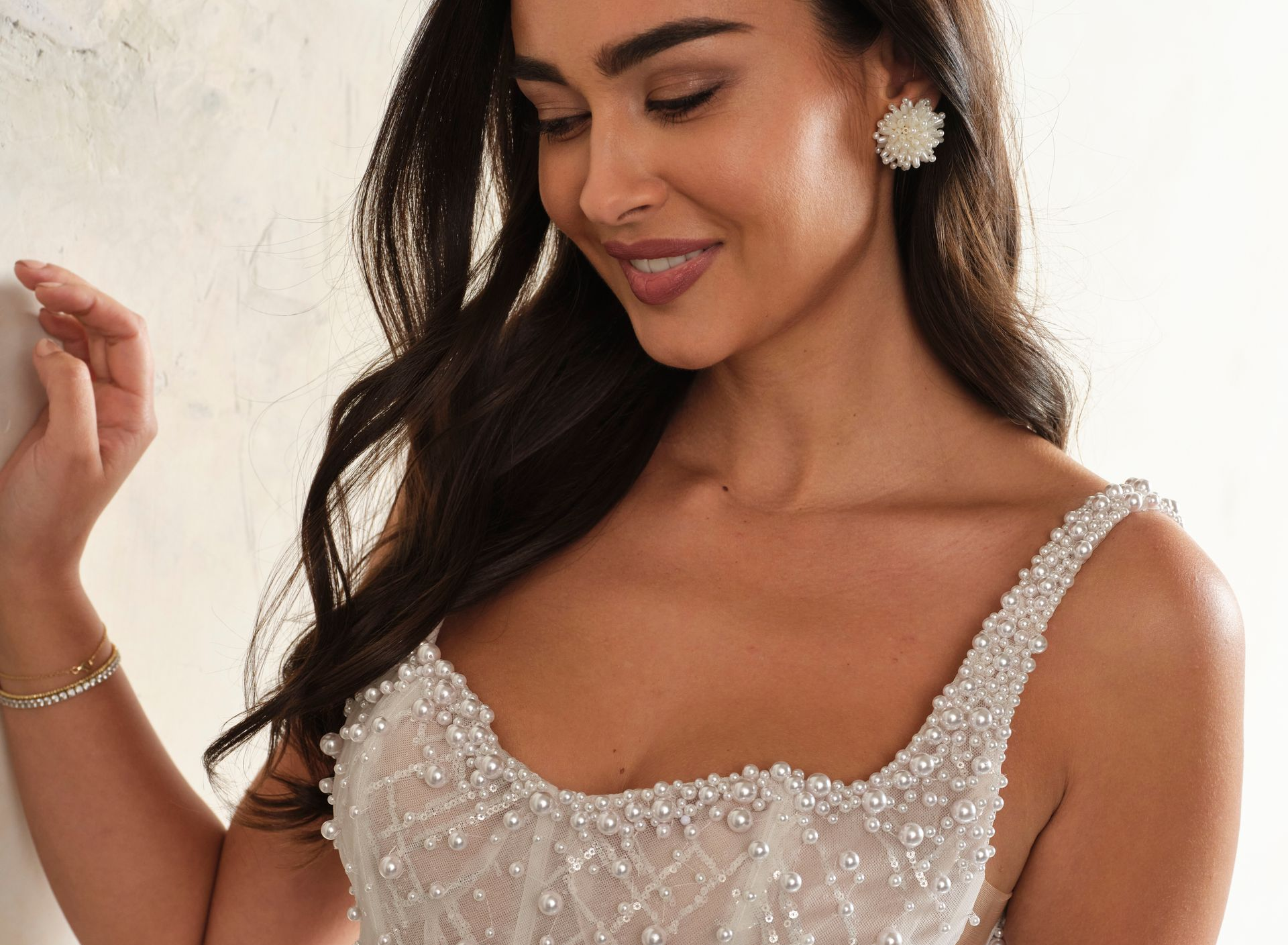Woman wearing a beaded wedding dress, smiling. Leaning against a white wall, wearing earrings.