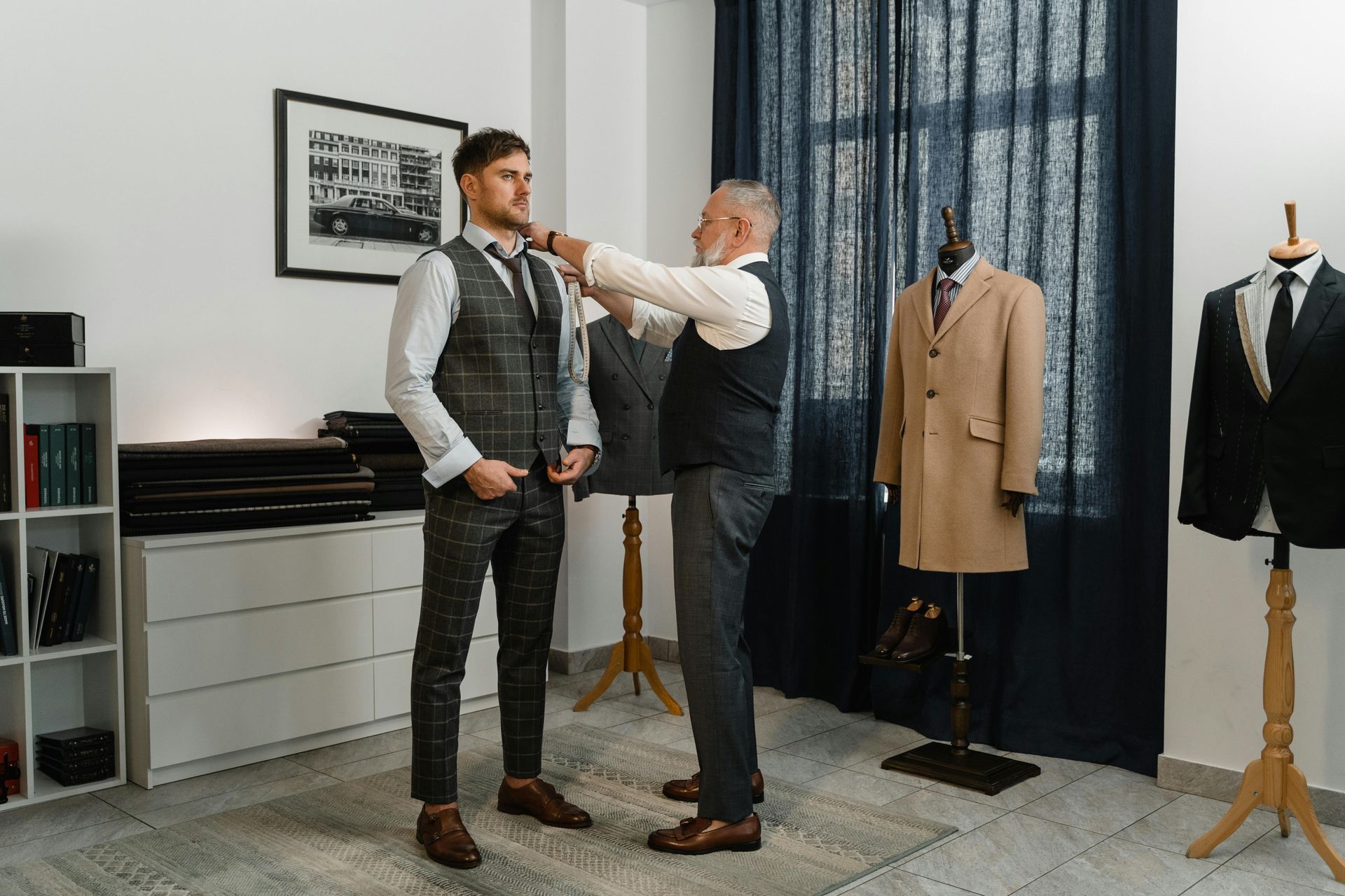 Tailor adjusting a man's tie in a suit shop. Mannequins with coats and suits, measuring tools visible.