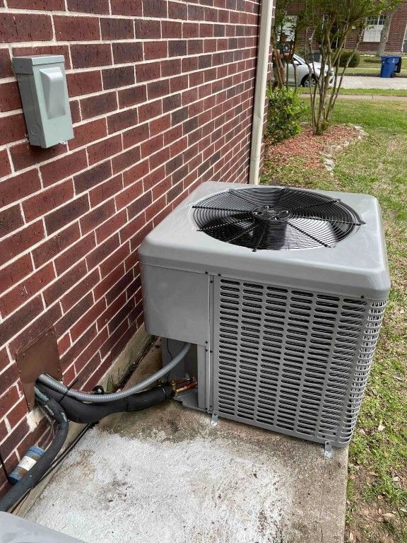 Outdoor air conditioning unit beside a brick house wall with exposed black hoses and a concrete pad.