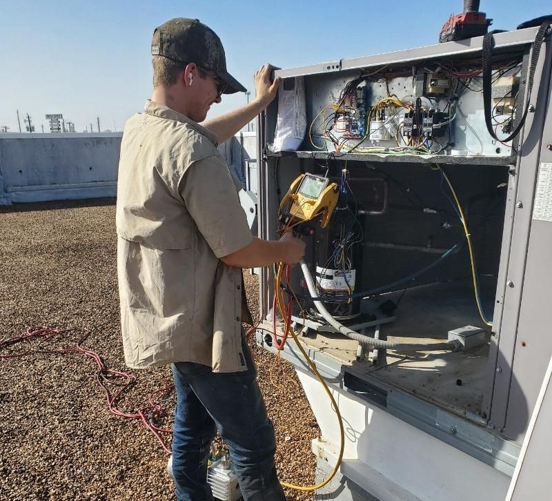 Technician inspecting rooftop electrical equipment with a multimeter beside an open control panel