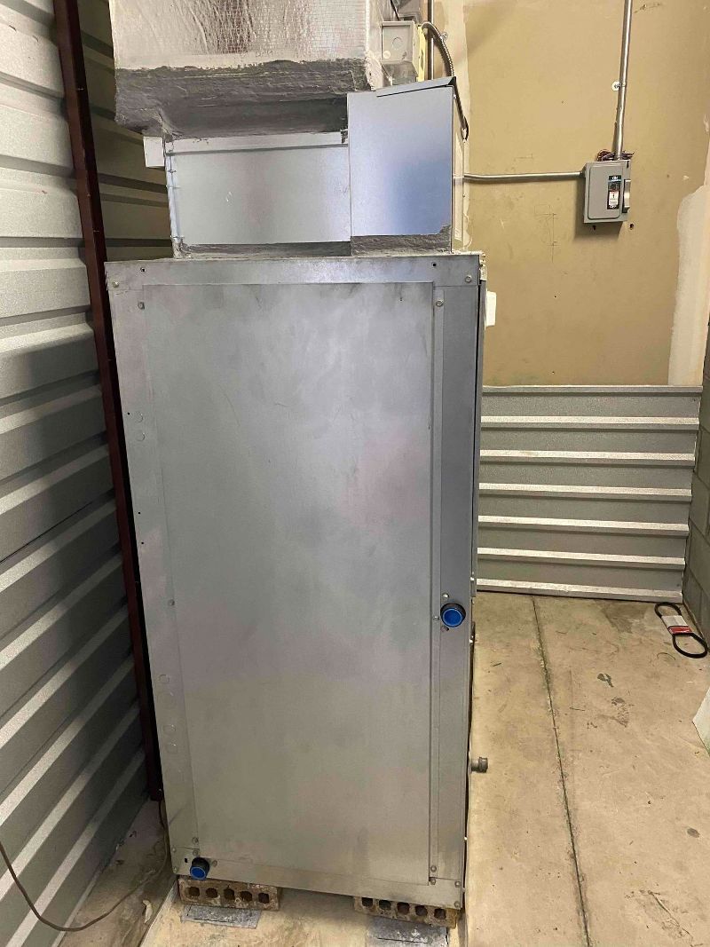 Tall stainless steel appliance in a basement or utility room beside louvered walls and exposed piping.