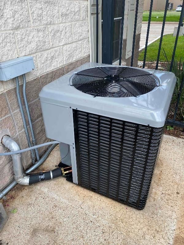 Outdoor air conditioning unit beside a brick wall and black metal fence on a concrete pad