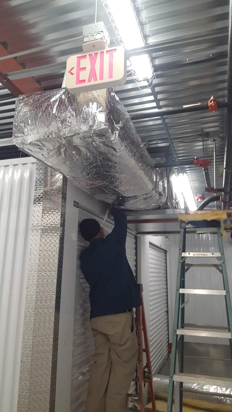 Worker installing silver ducting under an EXIT sign in a narrow industrial corridor