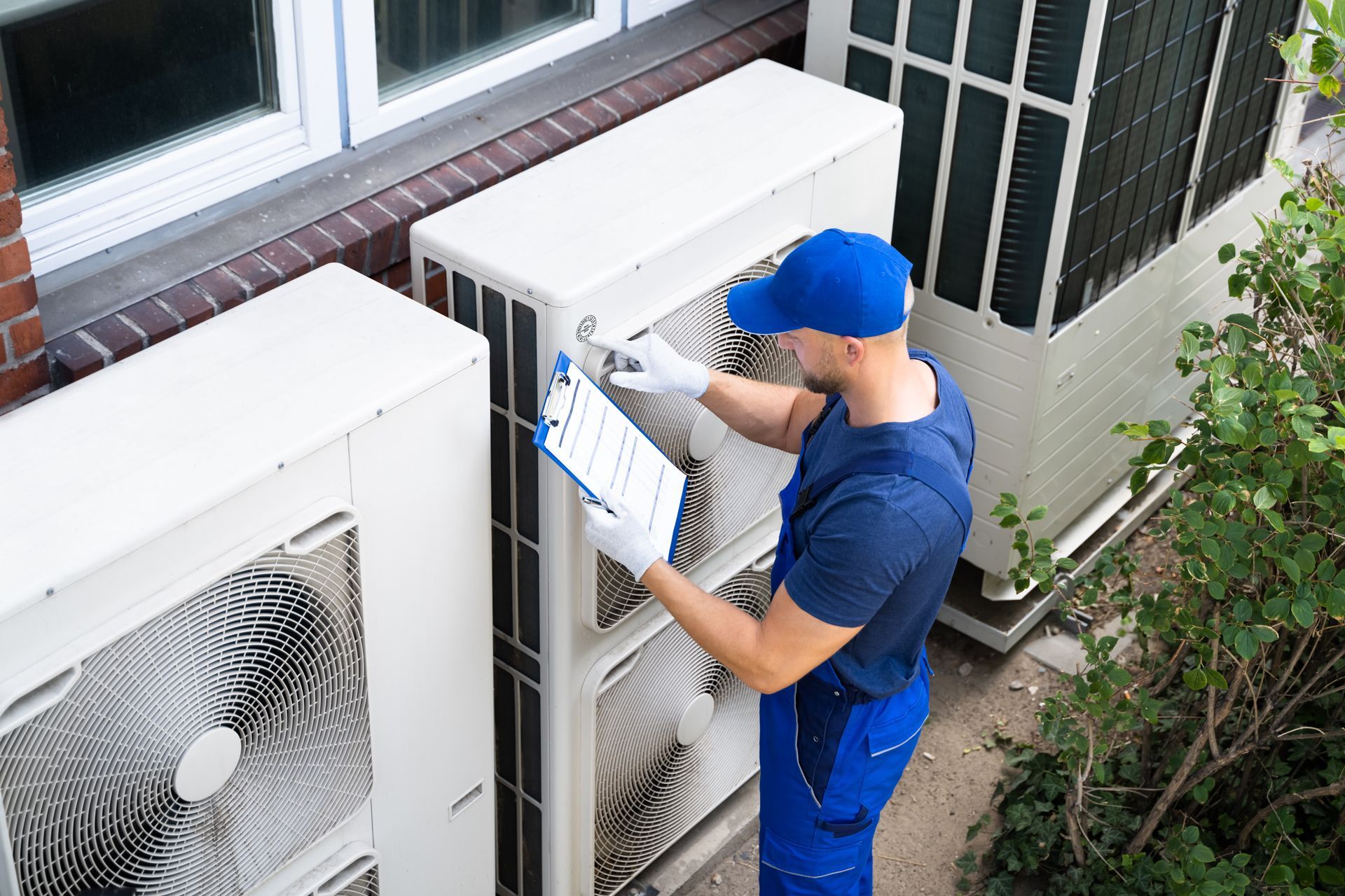 Technician in blue uniform inspecting an outdoor air conditioner unit with a clipboard.