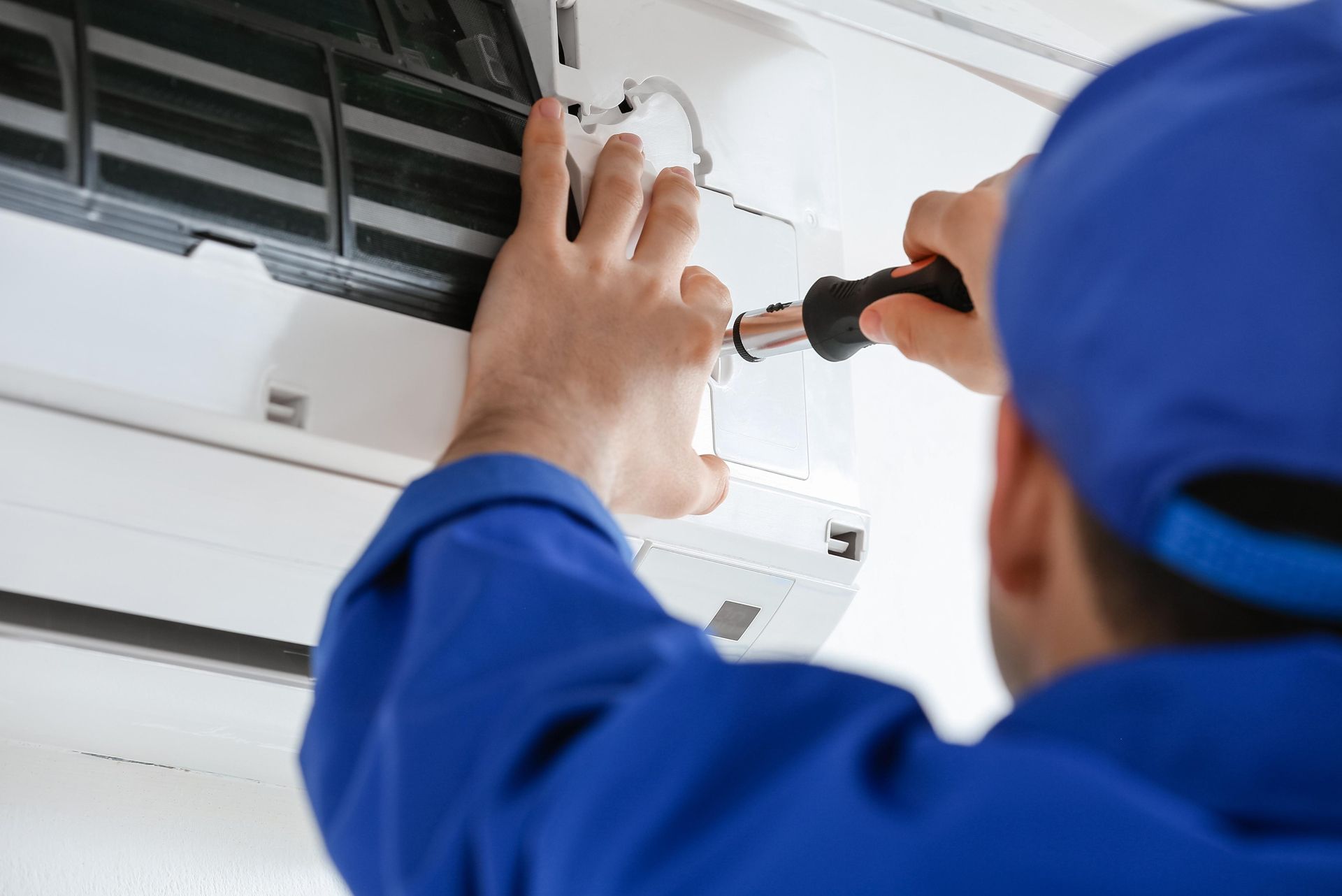 Technician in blue uniform repairing a wall-mounted air conditioner with a screwdriver