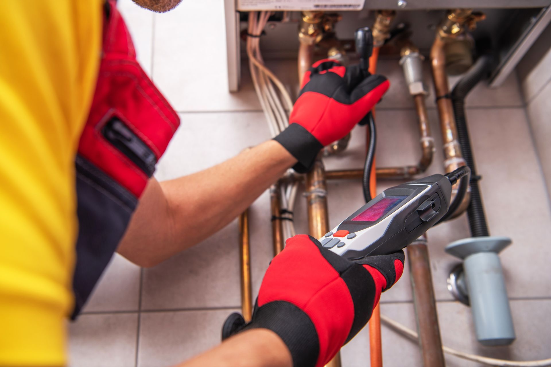 Technician in red gloves inspecting copper pipes under a sink with a wrench