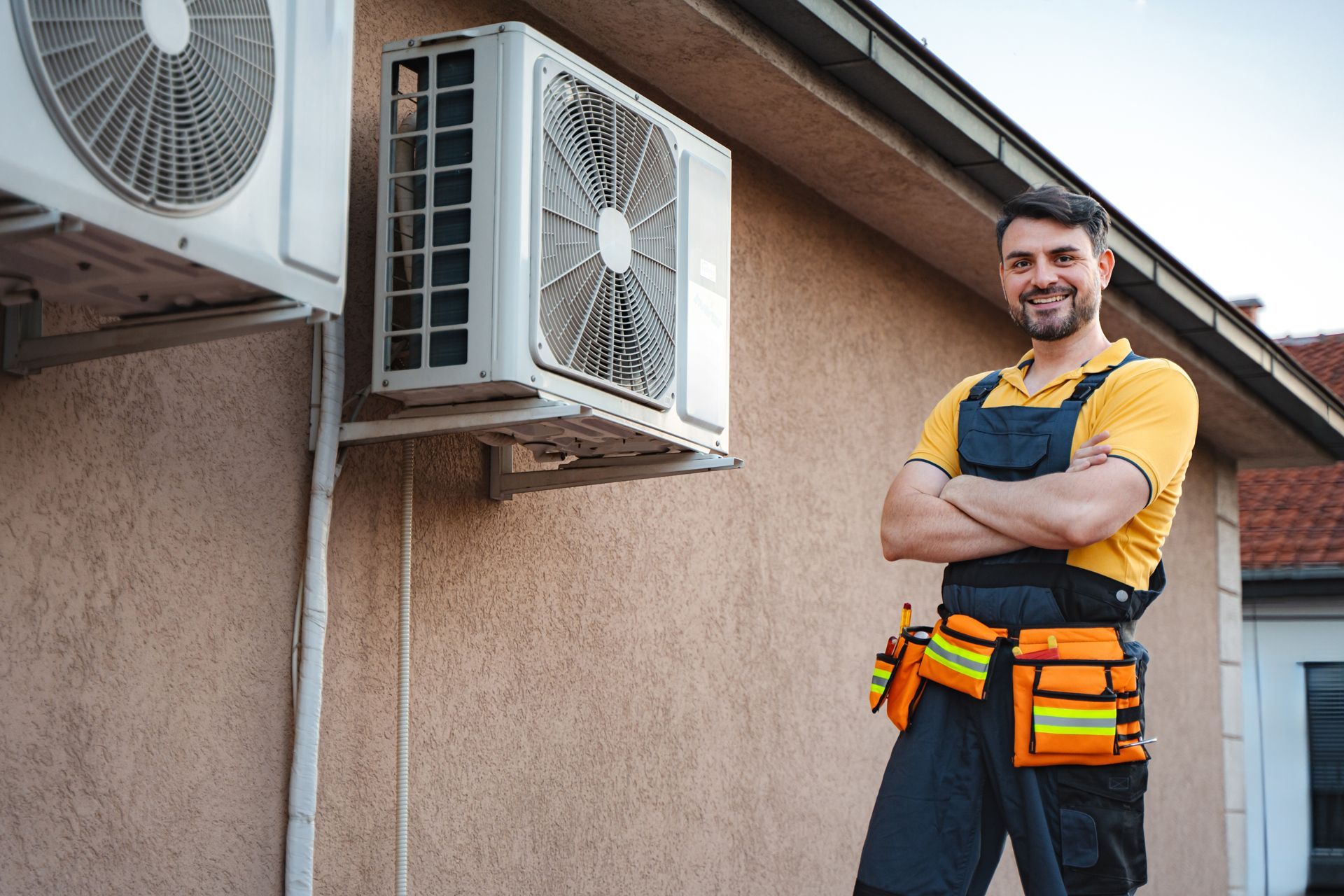 Technician with tool belt stands beside outdoor air conditioner units on a house exterior