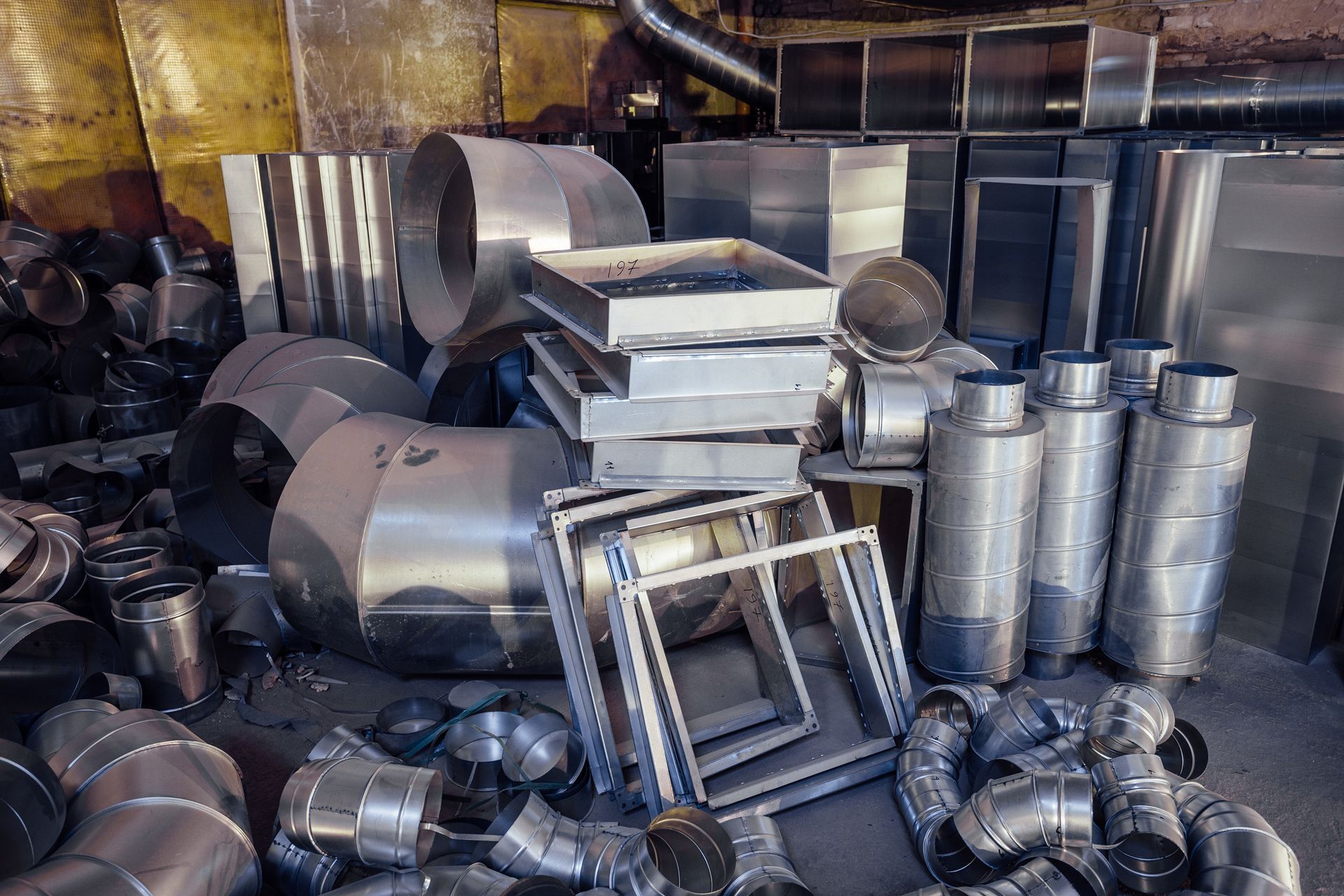 Stacked metal ducting and ventilation parts in a cluttered industrial storage area
