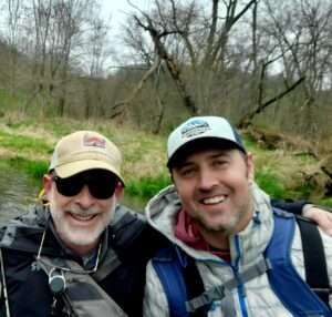 Two men smiling, wearing hats and sunglasses, in a wooded outdoor setting; one in a jacket, backpack, and fishing gear.