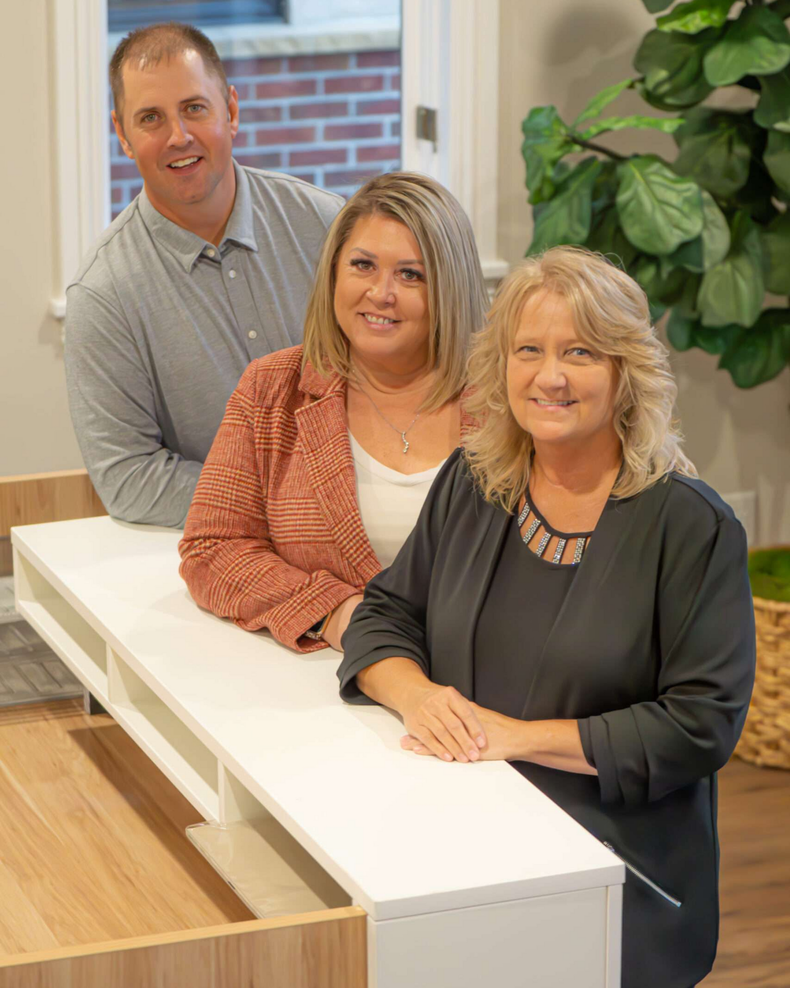Three people leaning on a white counter; two women and a man, smiling indoors.